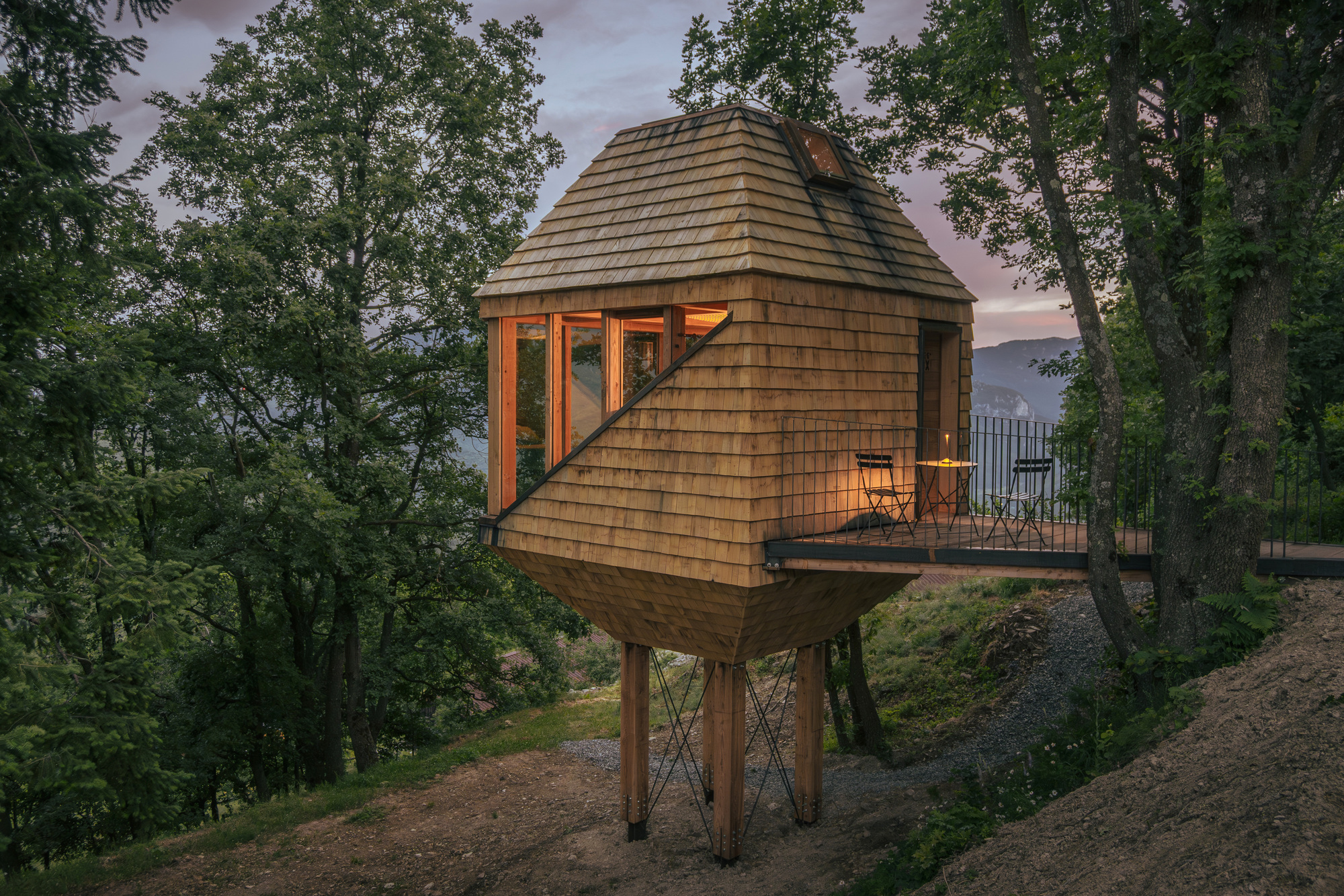Shingle-clad cabin on timber stilts with a railed deck and two chairs, glowing at dusk among trees on a slope
