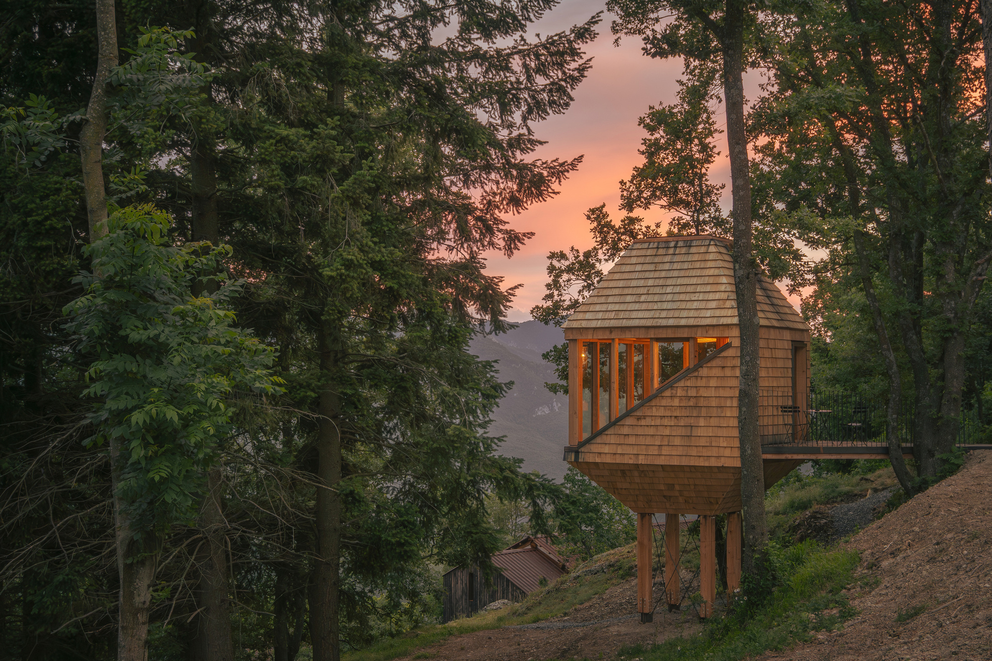 Shingled treehouse cabin raised on timber legs beside a trunk, with warm windows under an orange sunset sky