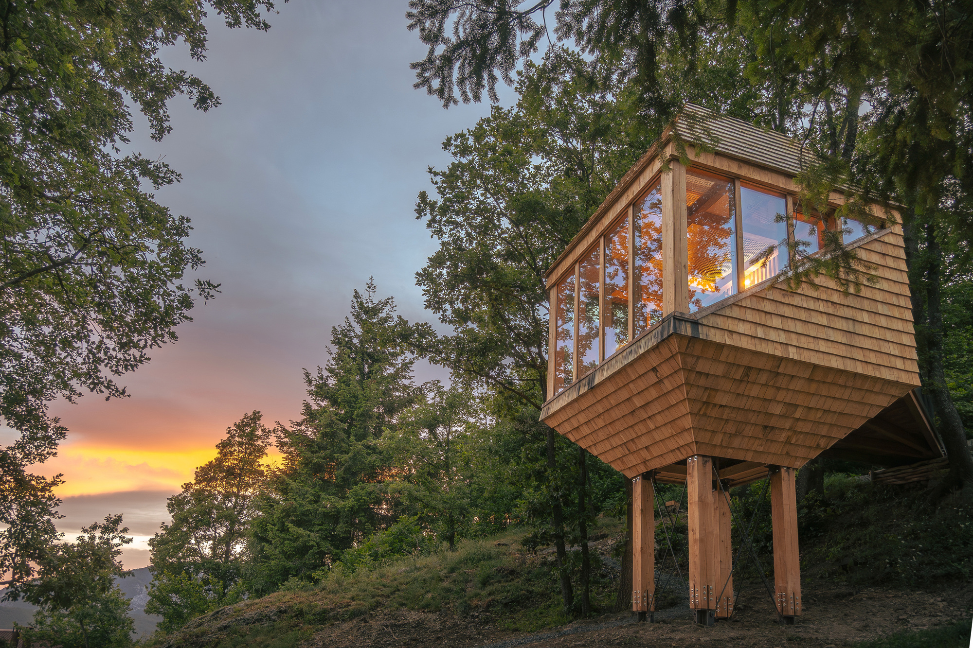 Low angle of the faceted shingled cabin on timber posts, its glazed side lit against a yellow and purple dusk sky