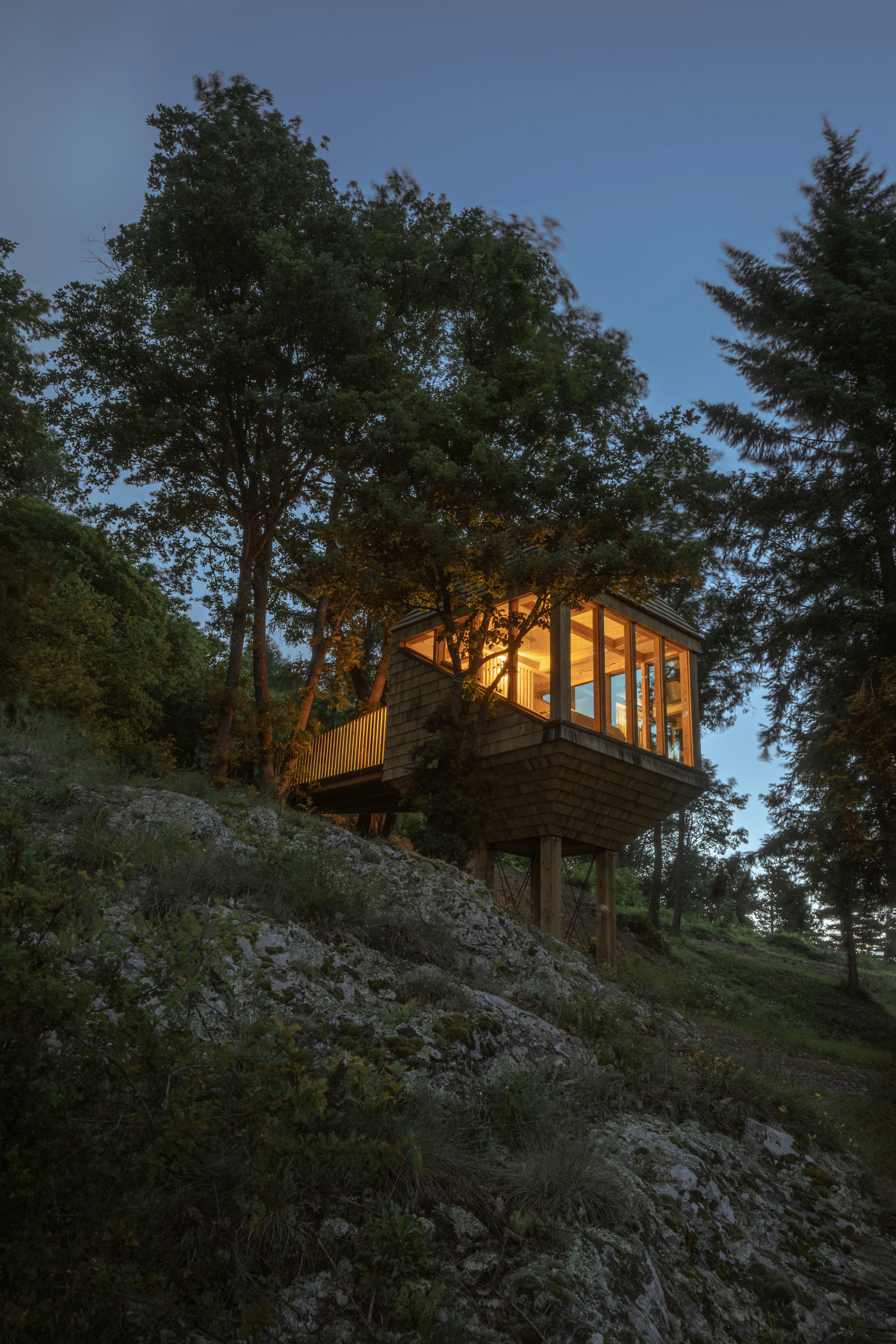 Shingle-clad cabin raised on timber posts above a rocky slope, lit from within under a deep blue evening sky