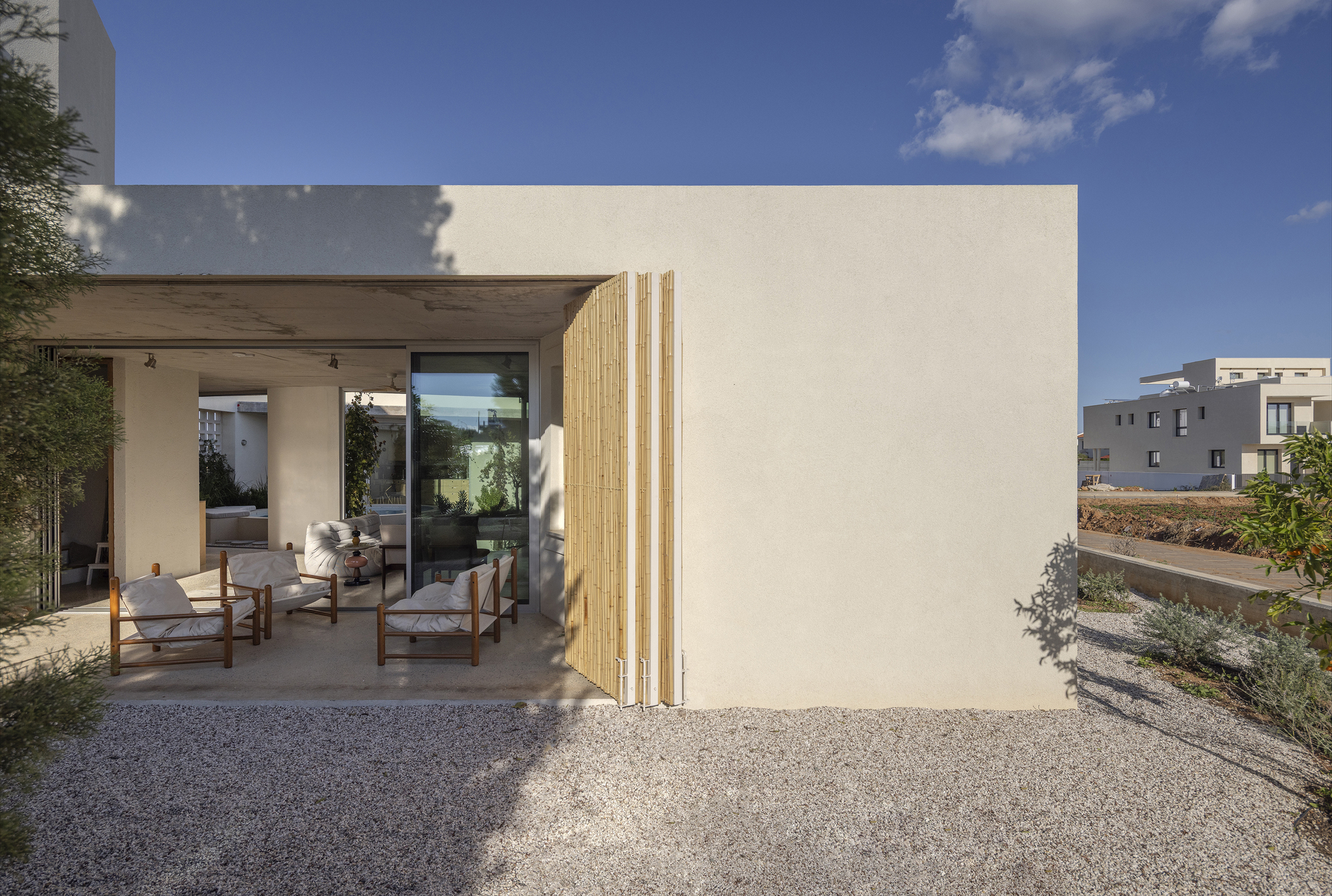 Covered terrace with wooden armchairs and a folding bamboo shutter beside a stucco wall, above a gravel garden in afternoon light