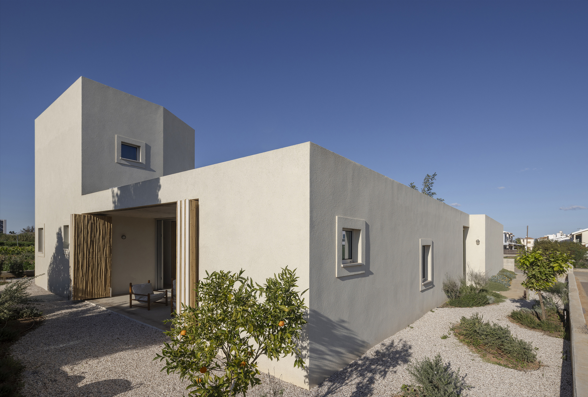 Corner view of the stucco house with square recessed windows, bamboo shutters and an orange tree in gravel at dusk