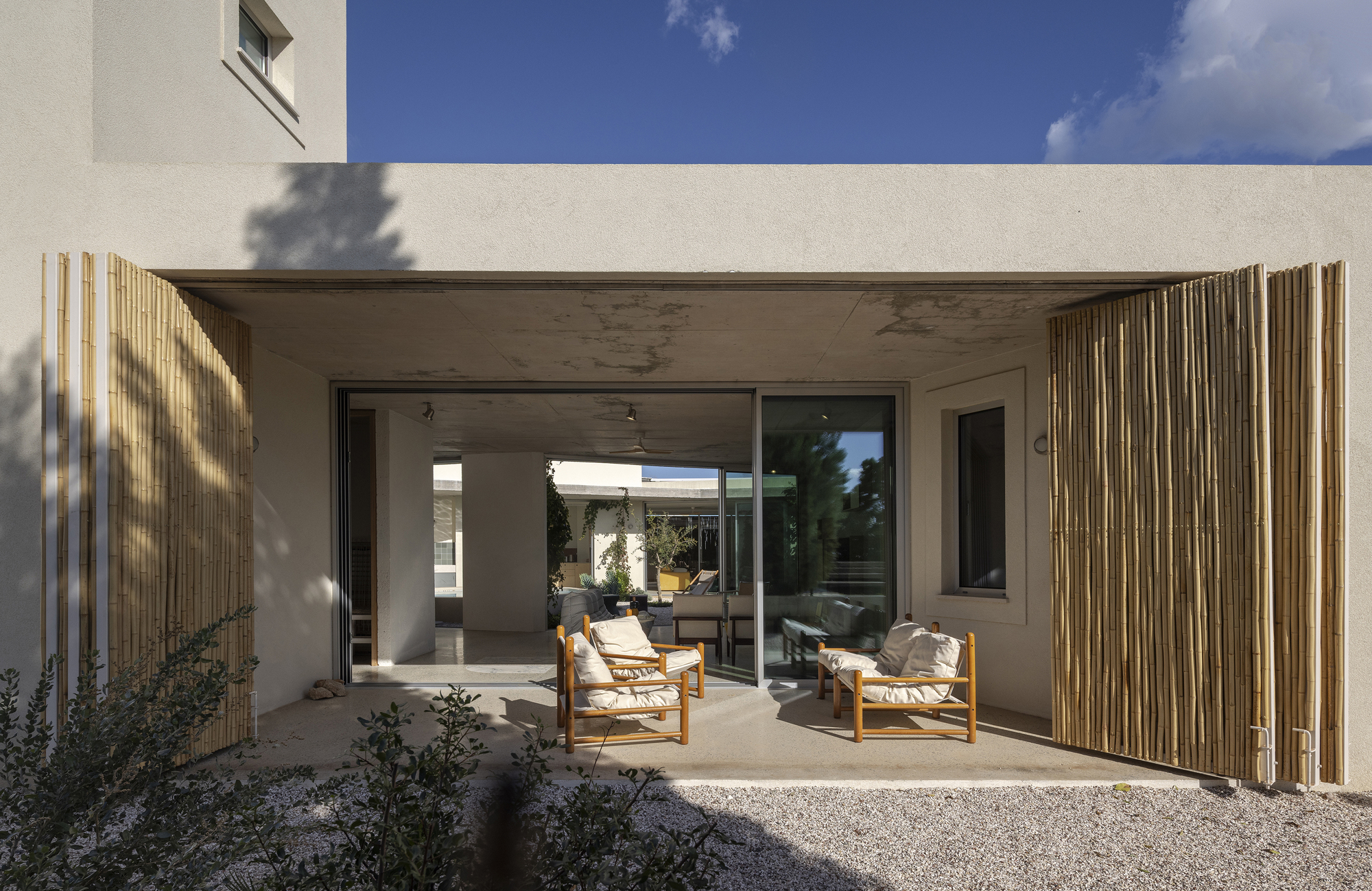 Covered terrace with folding bamboo screens, timber lounge chairs and a gravel yard under a clear blue sky
