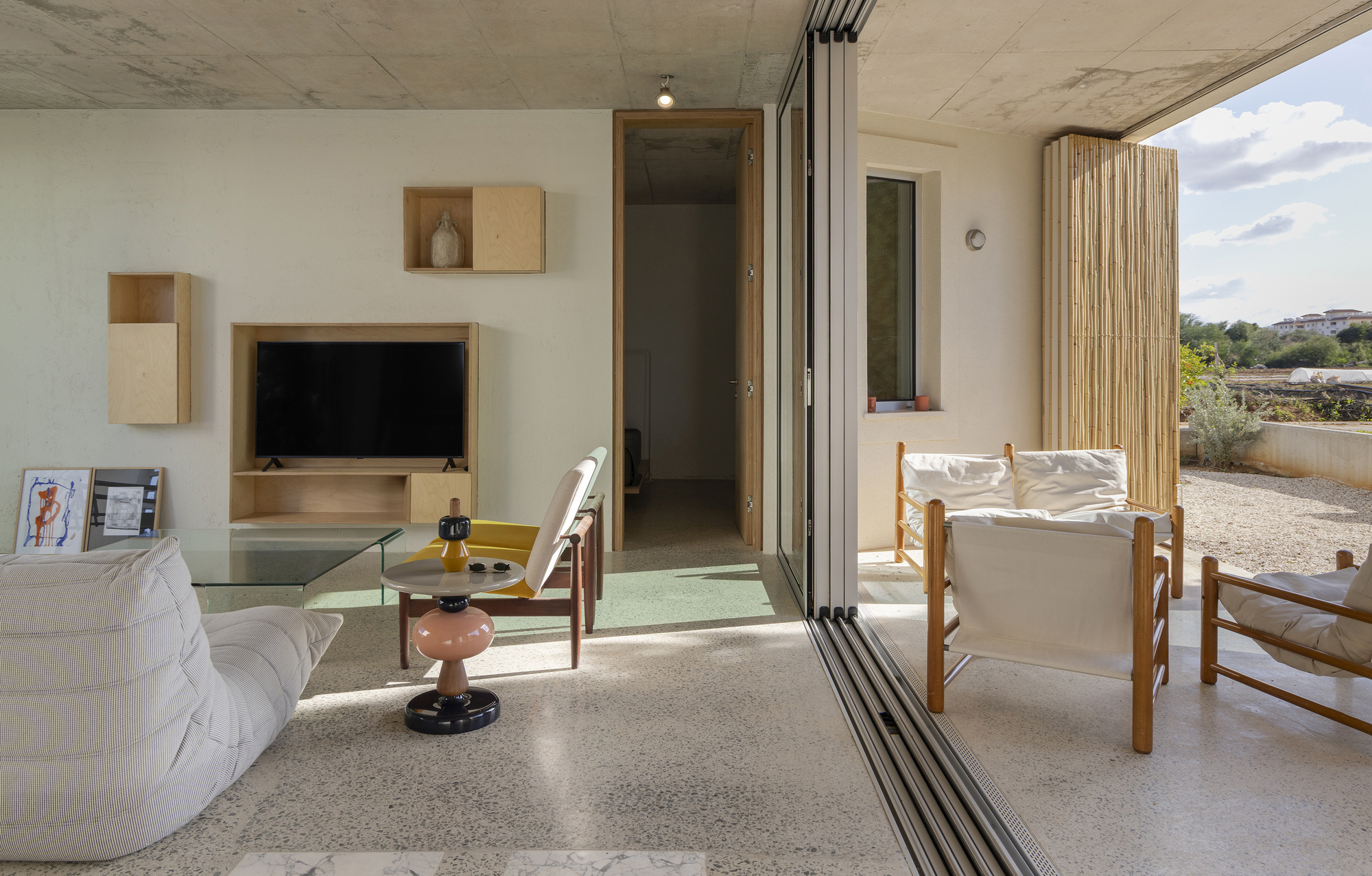 Living room with plywood shelving unit and a white lounger, opening through sliding glass to a gravel yard in afternoon sun