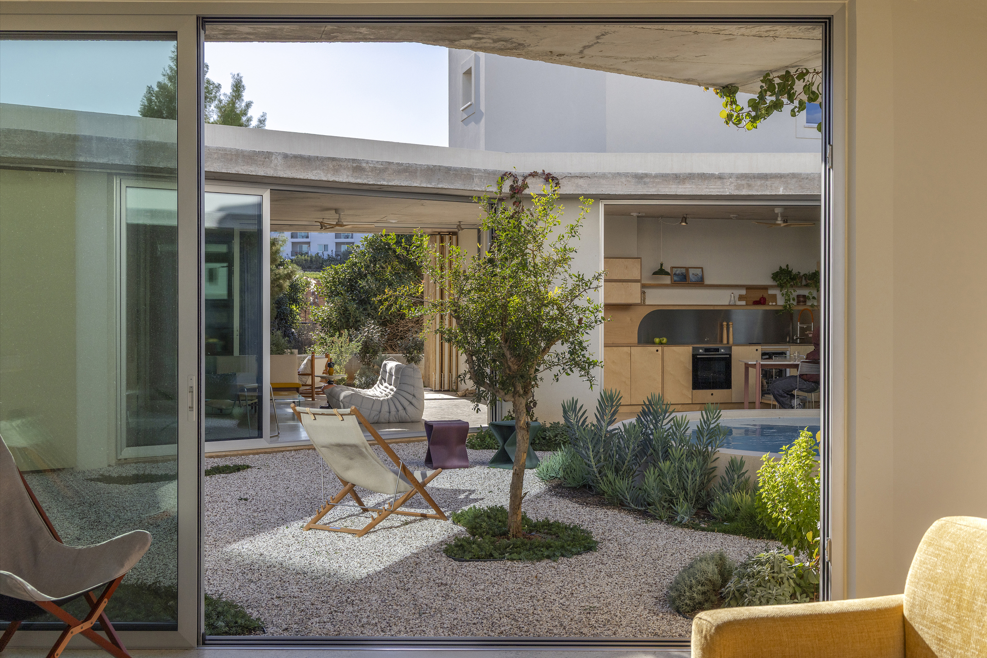 View through sliding glass doors to a gravel courtyard with a small tree, deck chair and a plywood kitchen beyond
