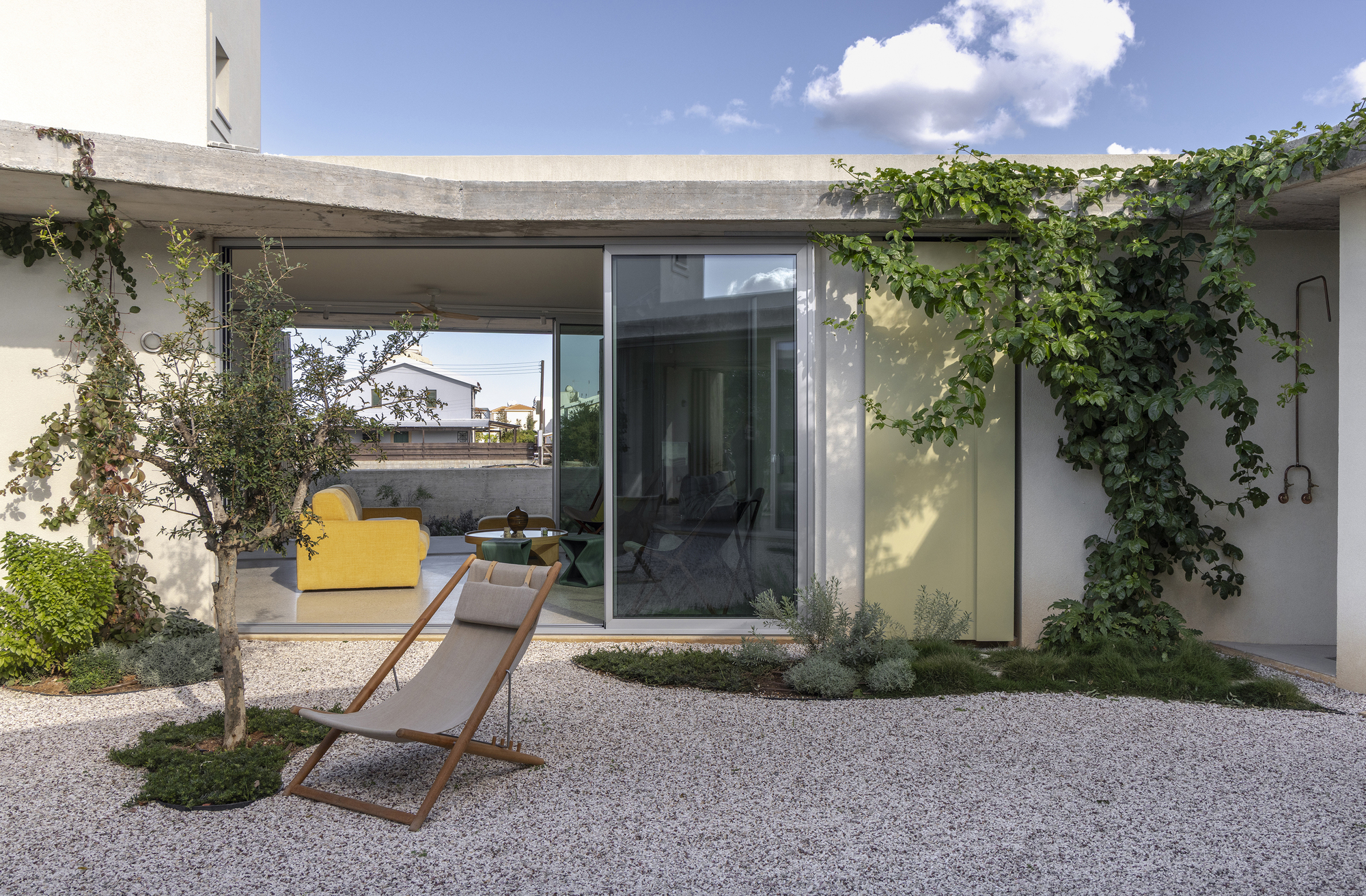 Courtyard facade with a climbing vine on the wall, a folding deck chair and a yellow armchair visible through the glass