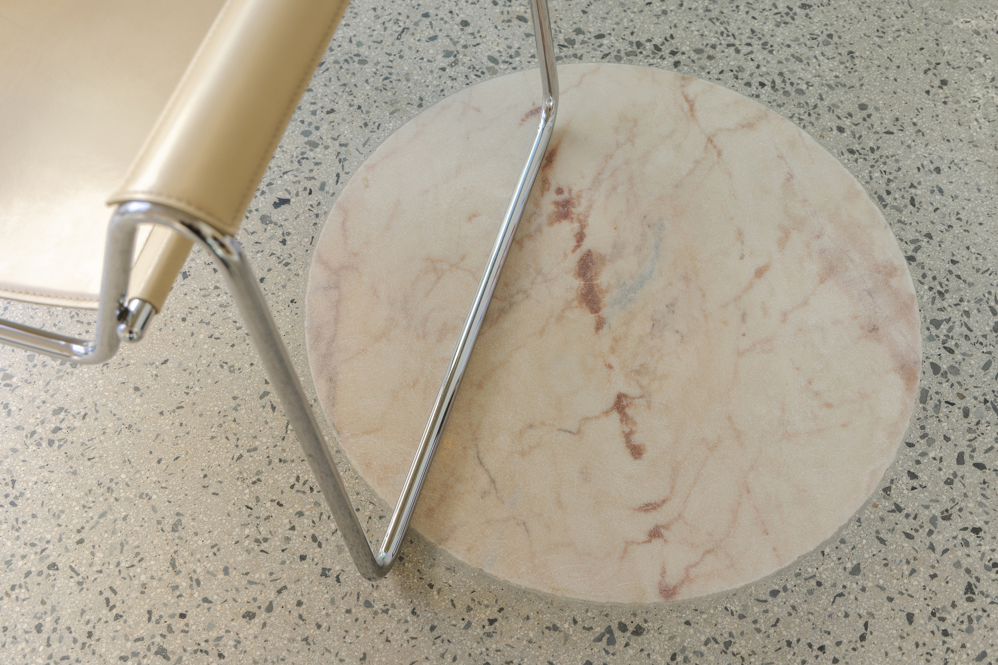 Top-down view of a chrome and leather chair leg beside a round pink marble inlay set in terrazzo
