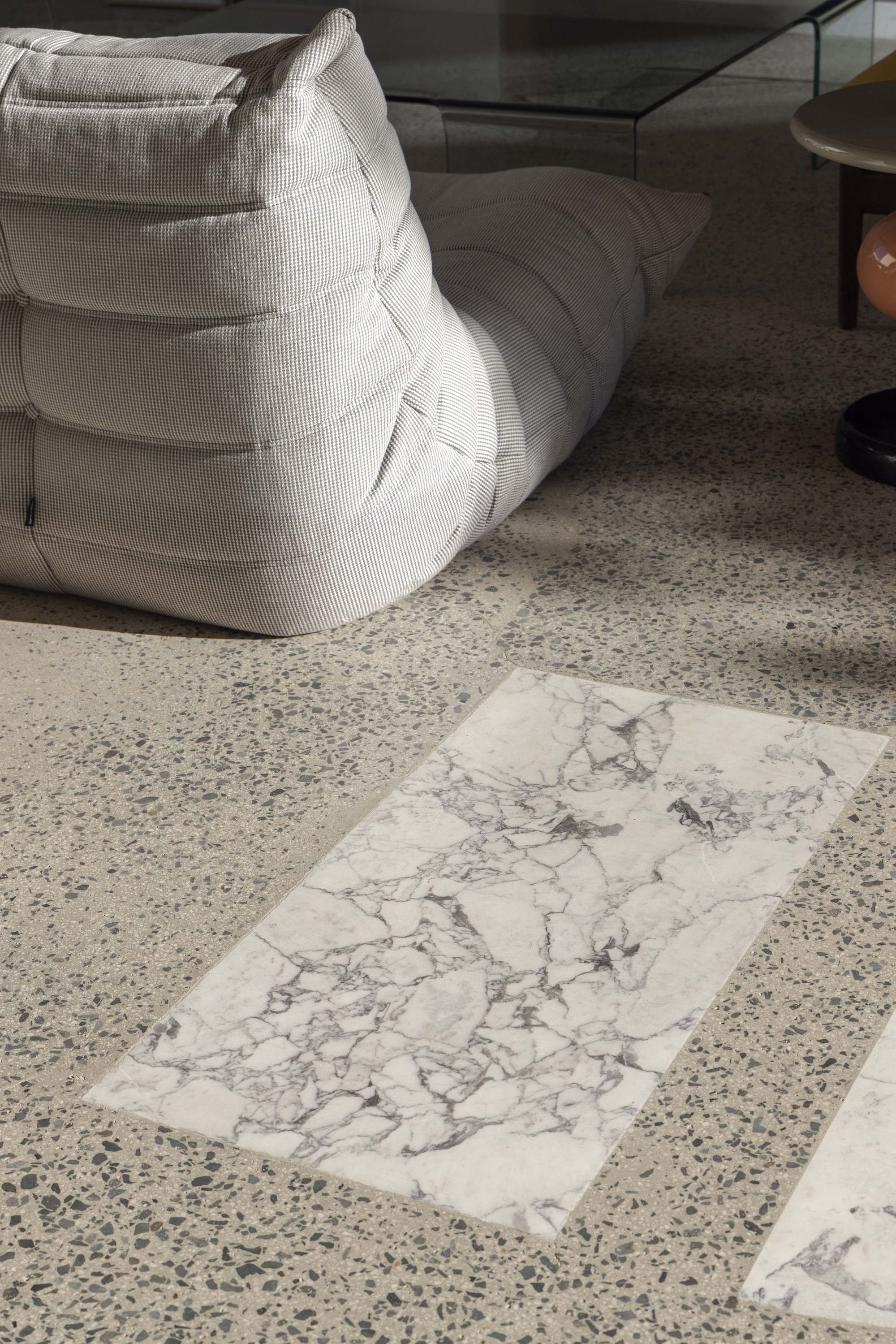 Rectangular white marble inlay in a terrazzo floor beside a grey fabric lounge seat in raking sunlight