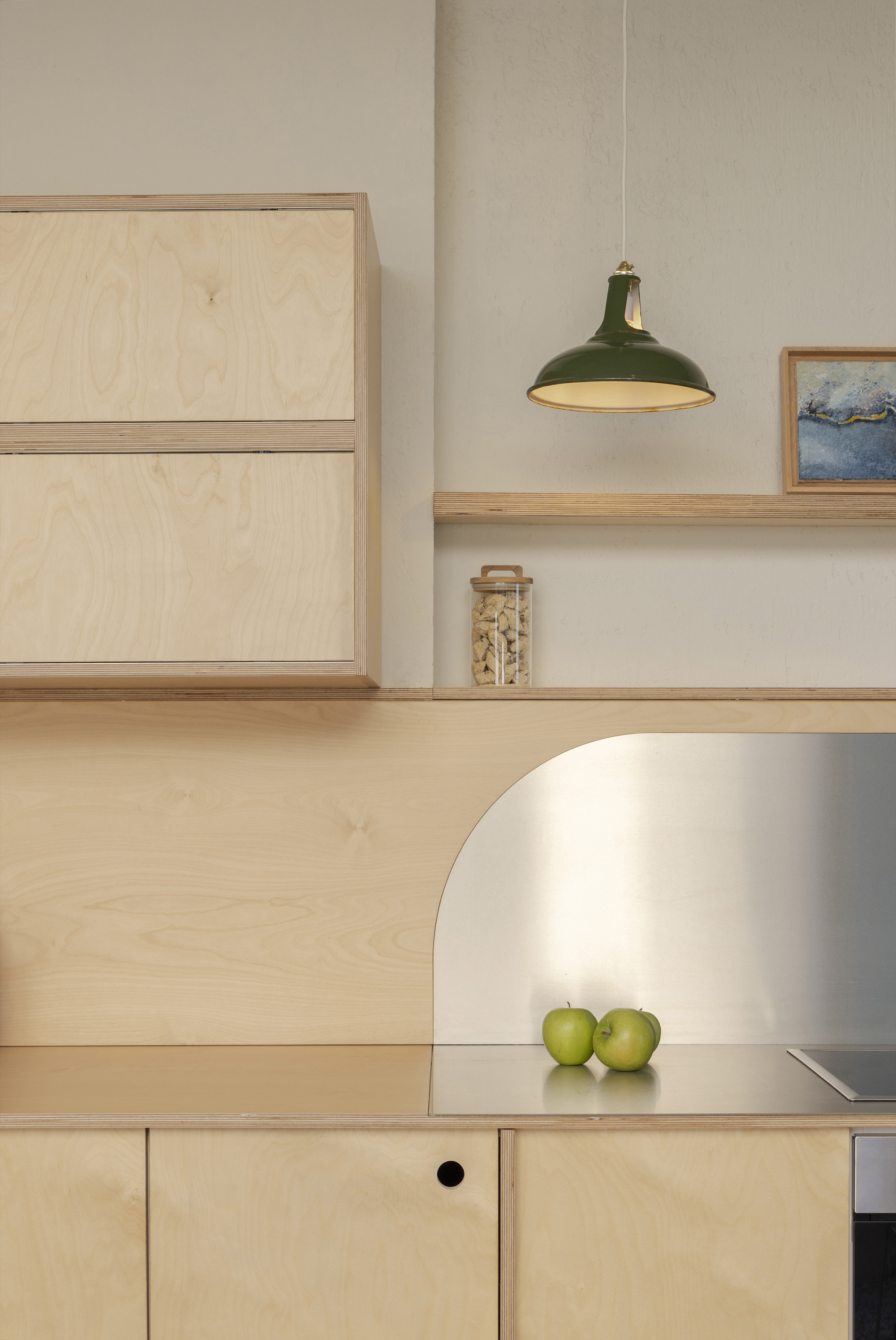 Plywood kitchen with an arched stainless splashback, green pendant lamp and two apples on the counter in soft daylight