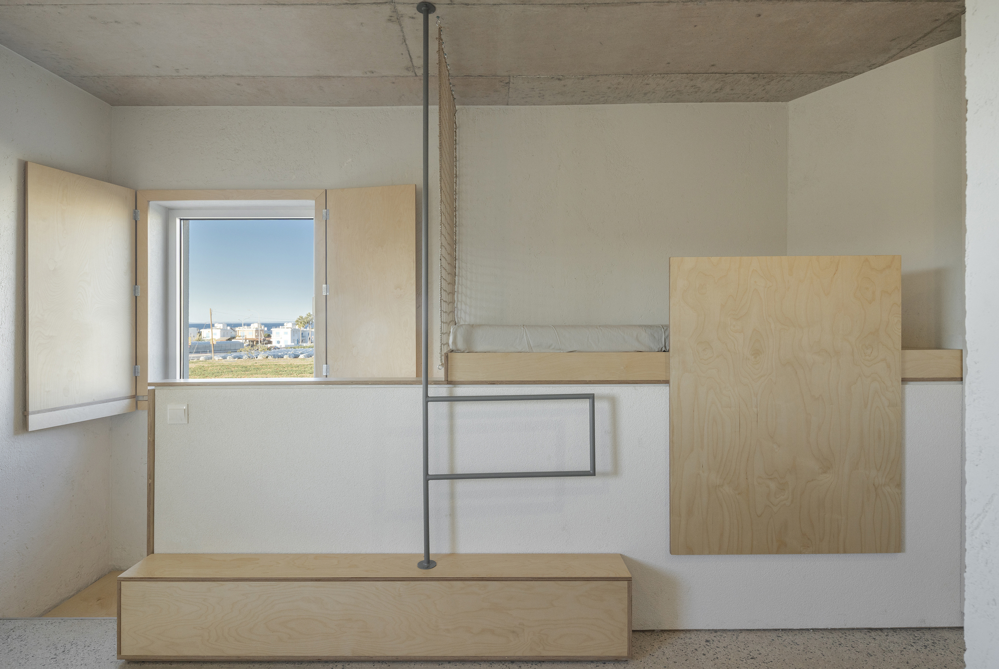 Plywood shutters, raised sleeping platform and steel pole with netting beneath a concrete ceiling, with a sunlit field through the window