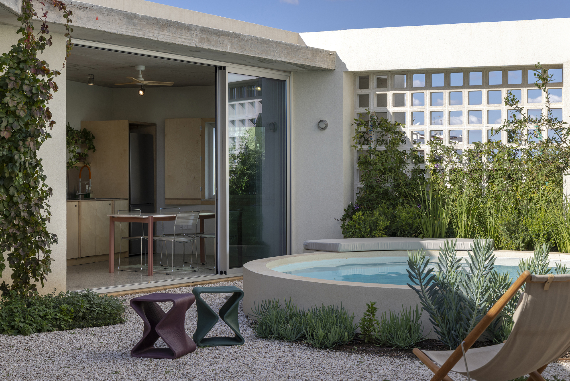 Round plunge pool beside an open sliding glass door, with a perforated screen wall, gravel court and folding canvas chair in daylight