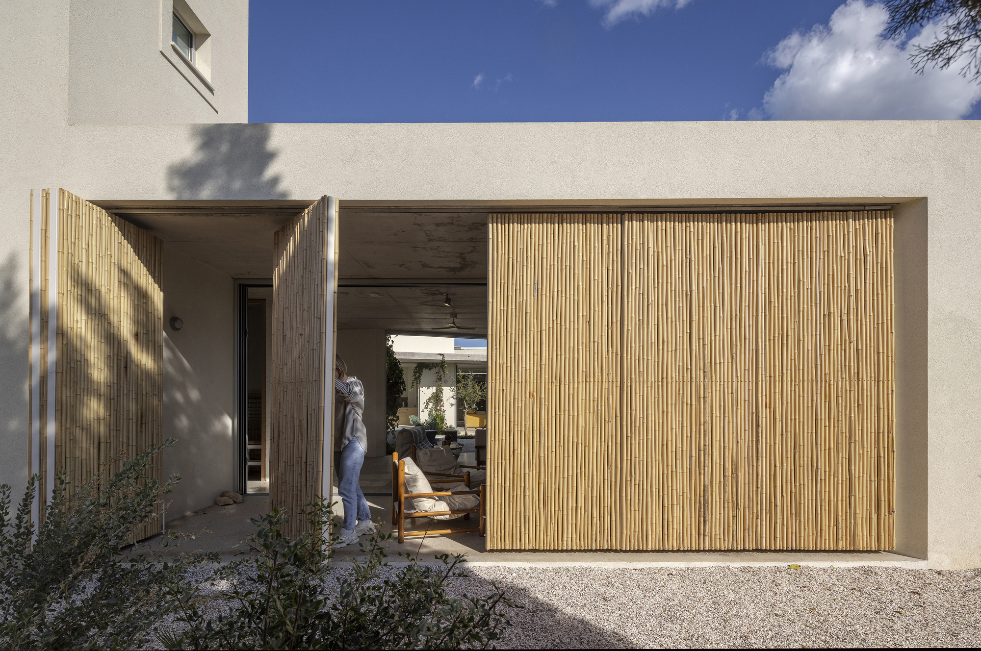 Bamboo sliding shutters pulled partly open on a plastered wall, with a person standing at the threshold under a blue sky