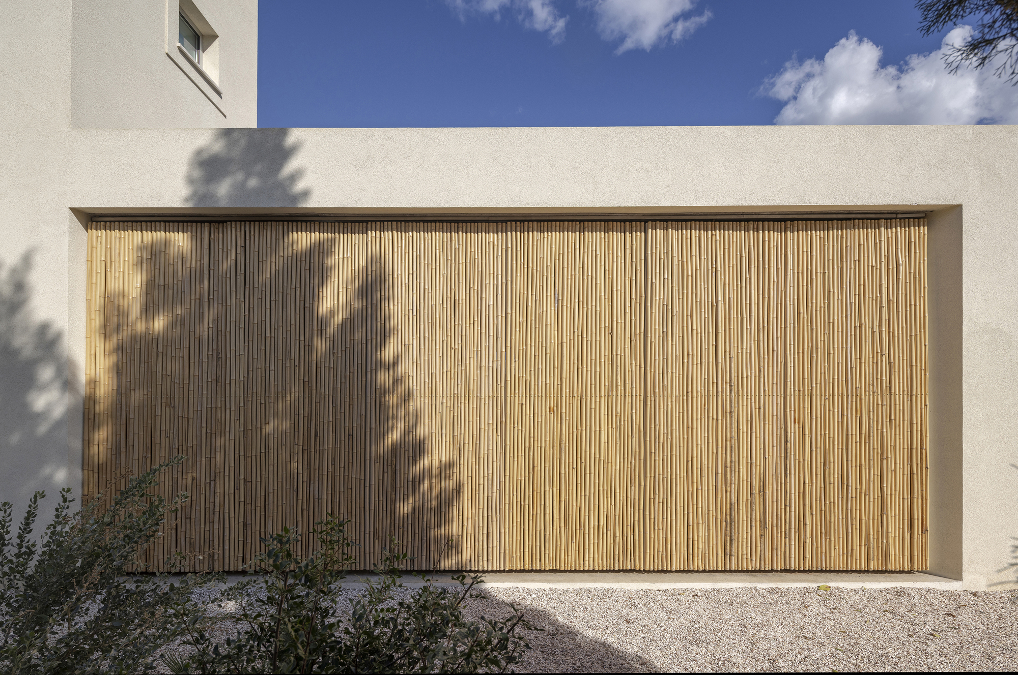 Closed bamboo shutter panel set into a plain plastered facade, with tree shadows falling across the cane in afternoon sun