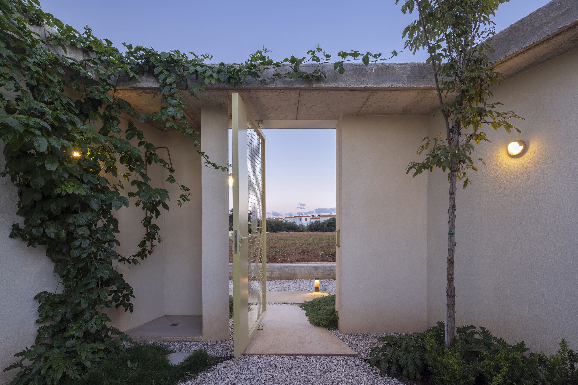 Entrance gate pivoted open beneath a concrete canopy, with a vine on the wall and a path lit at dusk