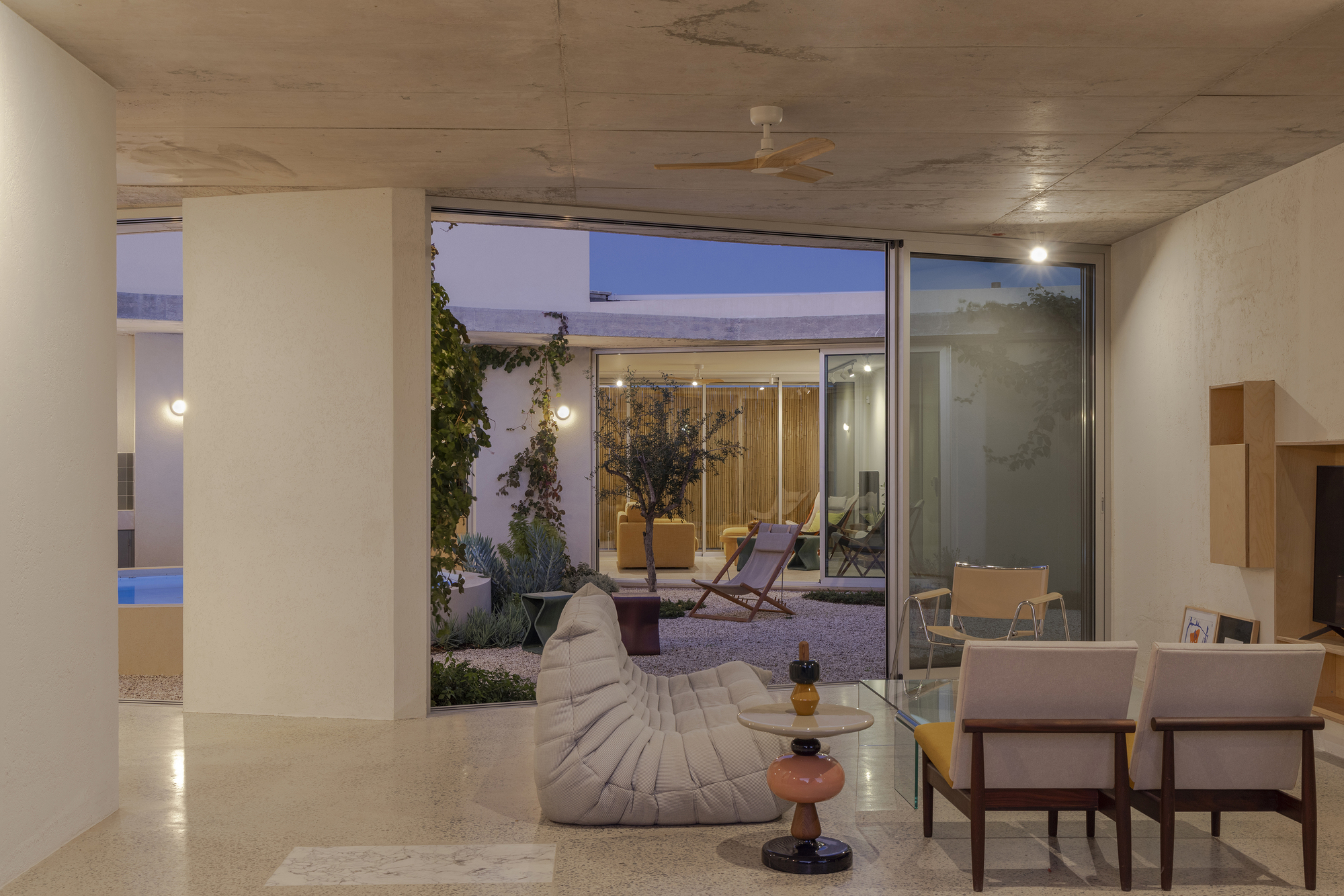 Living room with a low sofa and lounge chairs facing a gravel courtyard with an olive tree, seen through sliding glass at dusk