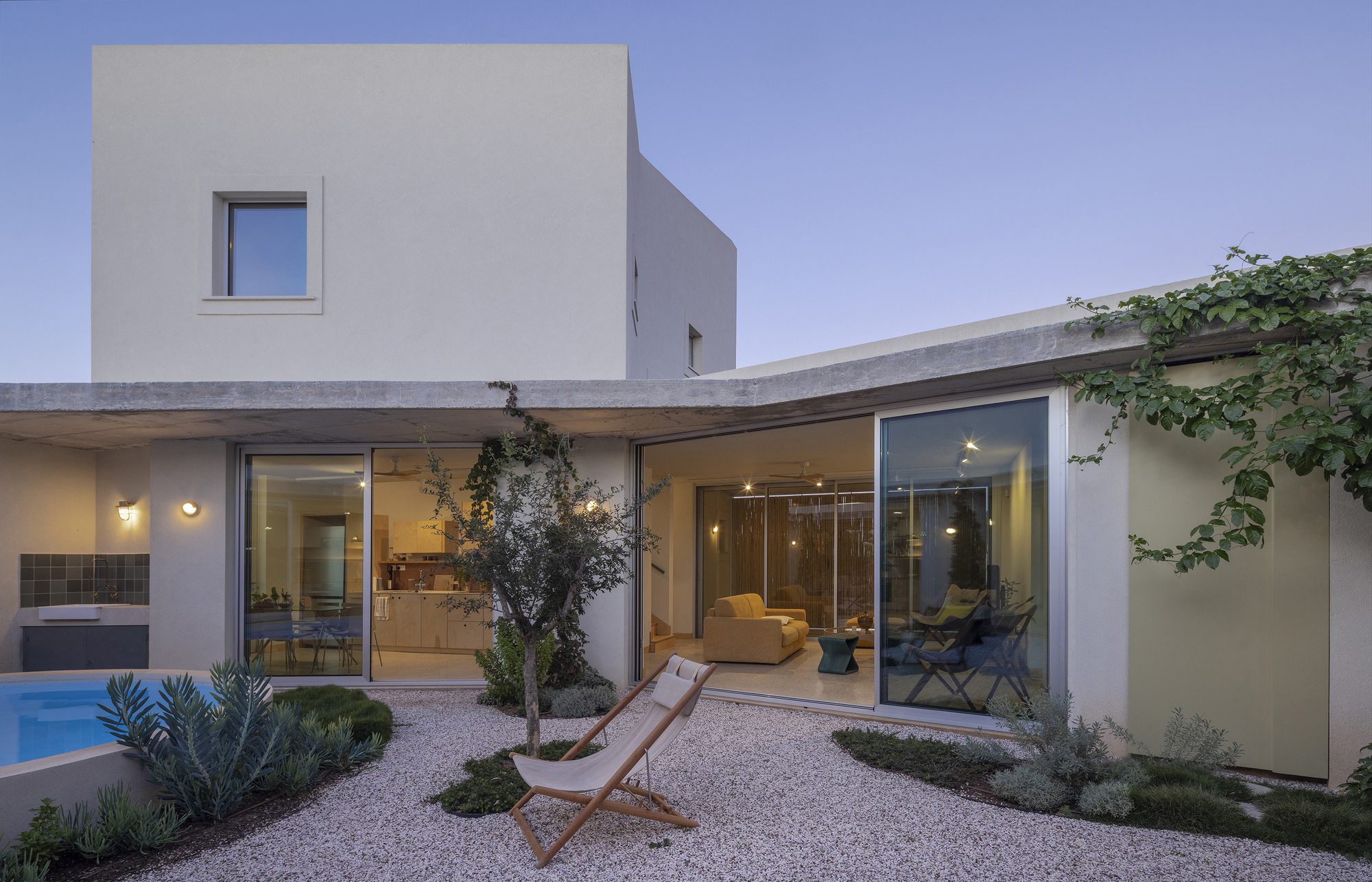 Courtyard elevation with a white upper volume, a thin concrete roof edge and glazed walls above a gravel garden with a canvas chair at dusk