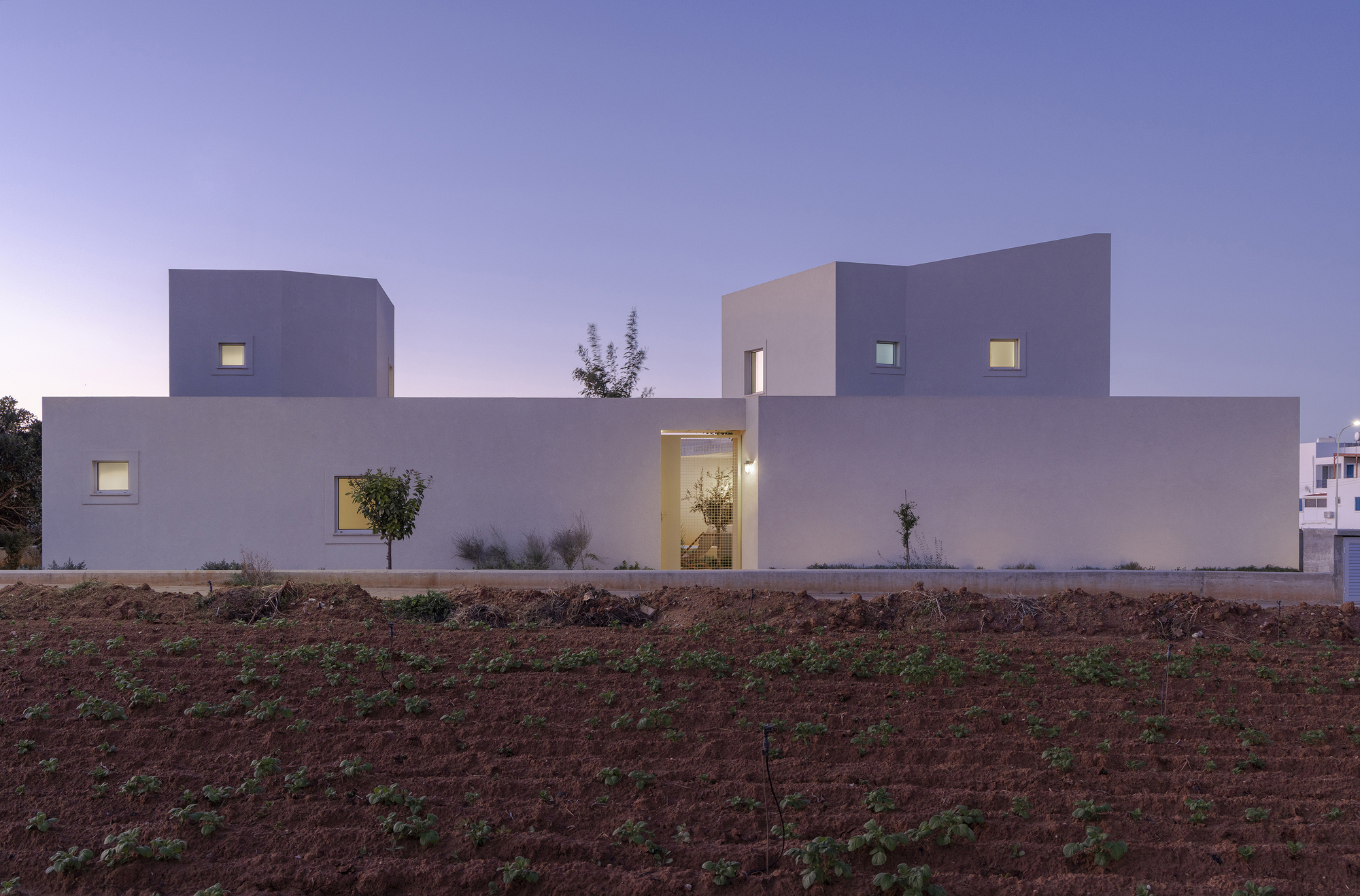 Low white-rendered house with two raised blocks and a lit entrance opening, seen across a plowed field at dusk