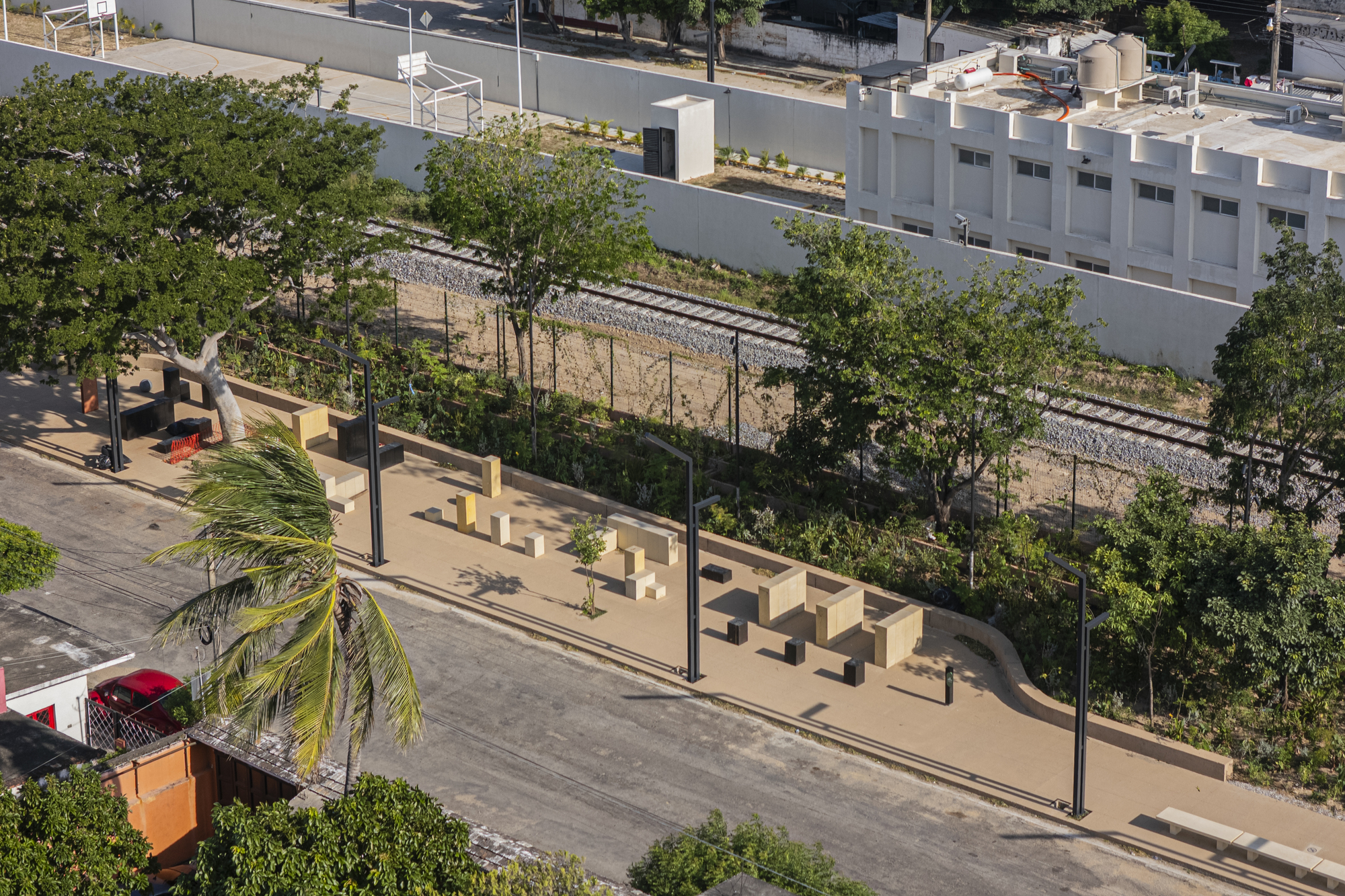 Aerial view of a sand-colored linear plaza with scattered block seating and black lamp posts beside a railway track and tree canopy