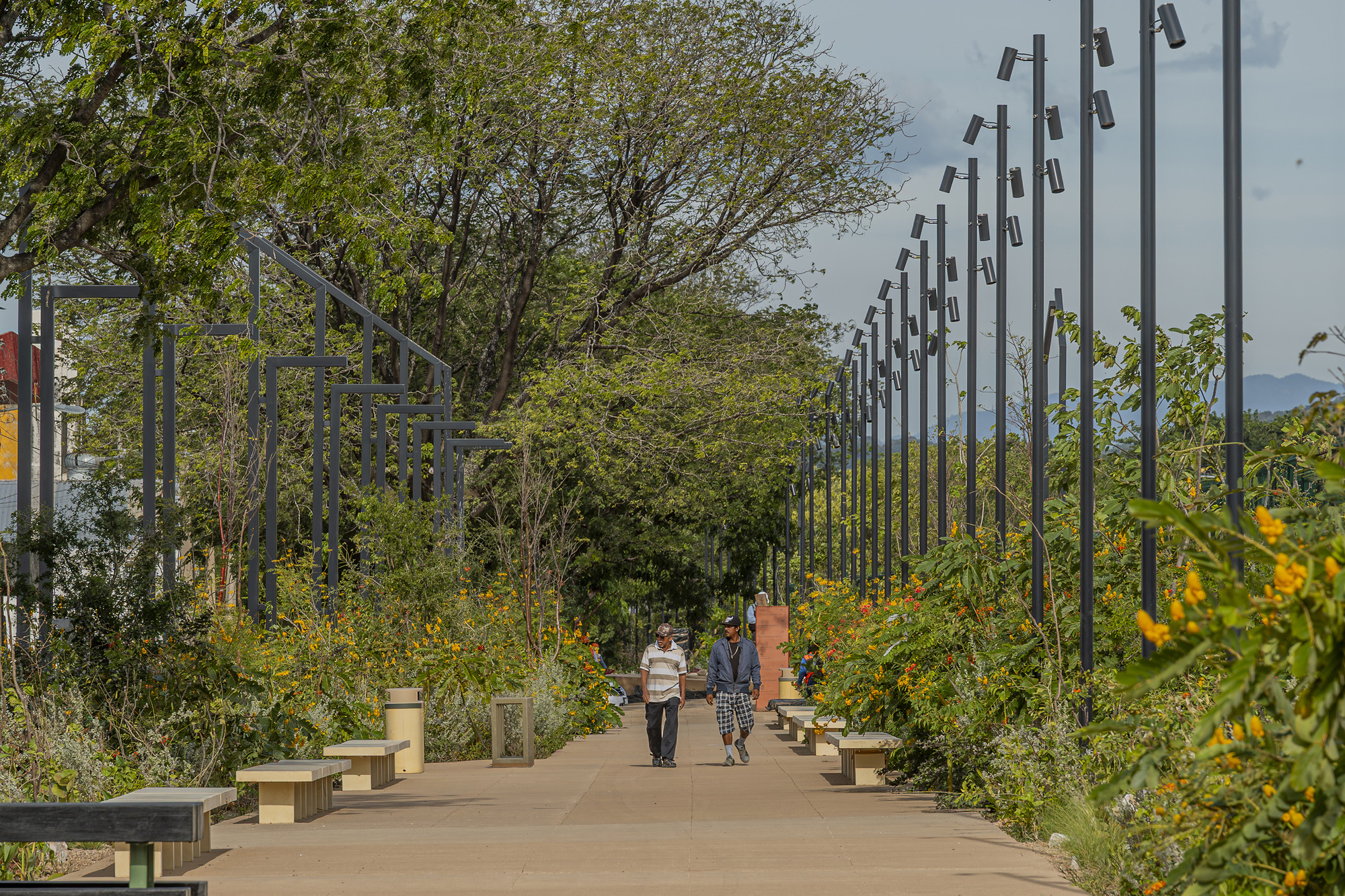 Two men walking along a wide paved promenade lined with benches, tall black light poles and flowering yellow shrubs under a hazy sky