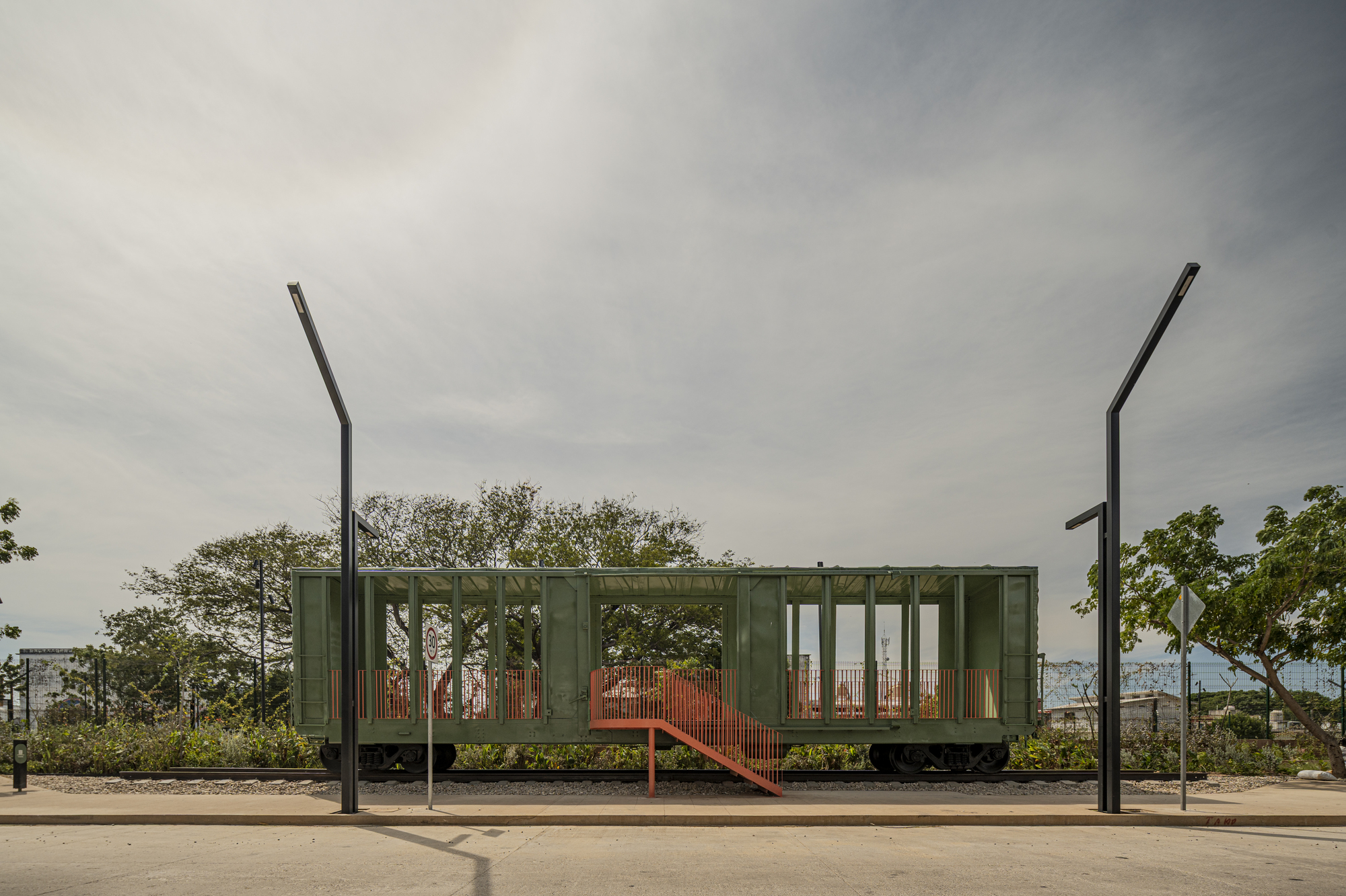 Green-painted freight car with orange stair and railings, set on tracks between two black lamp posts under an overcast sky