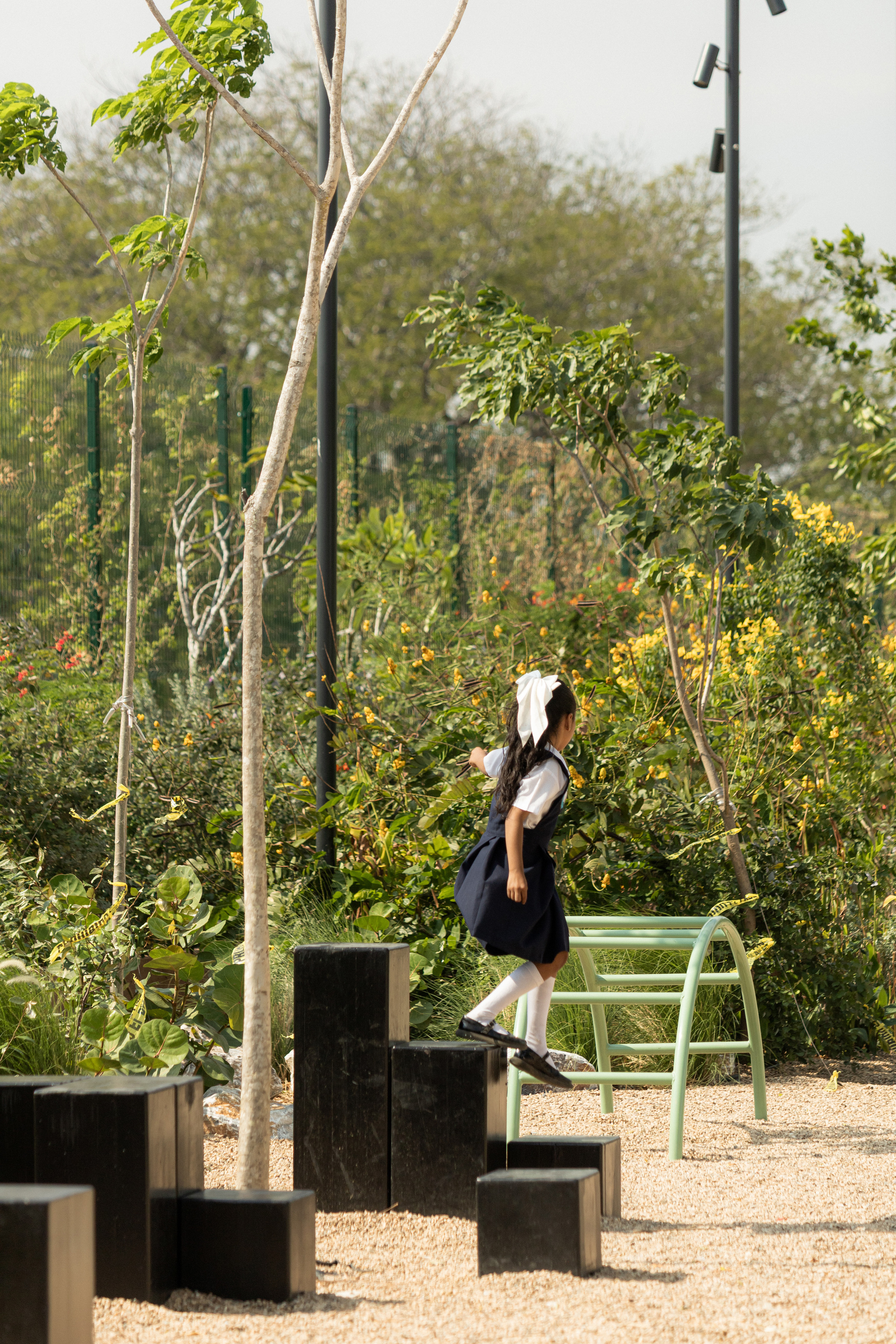 Girl in school uniform stepping across black timber blocks on a gravel play area among flowering shrubs and young trees