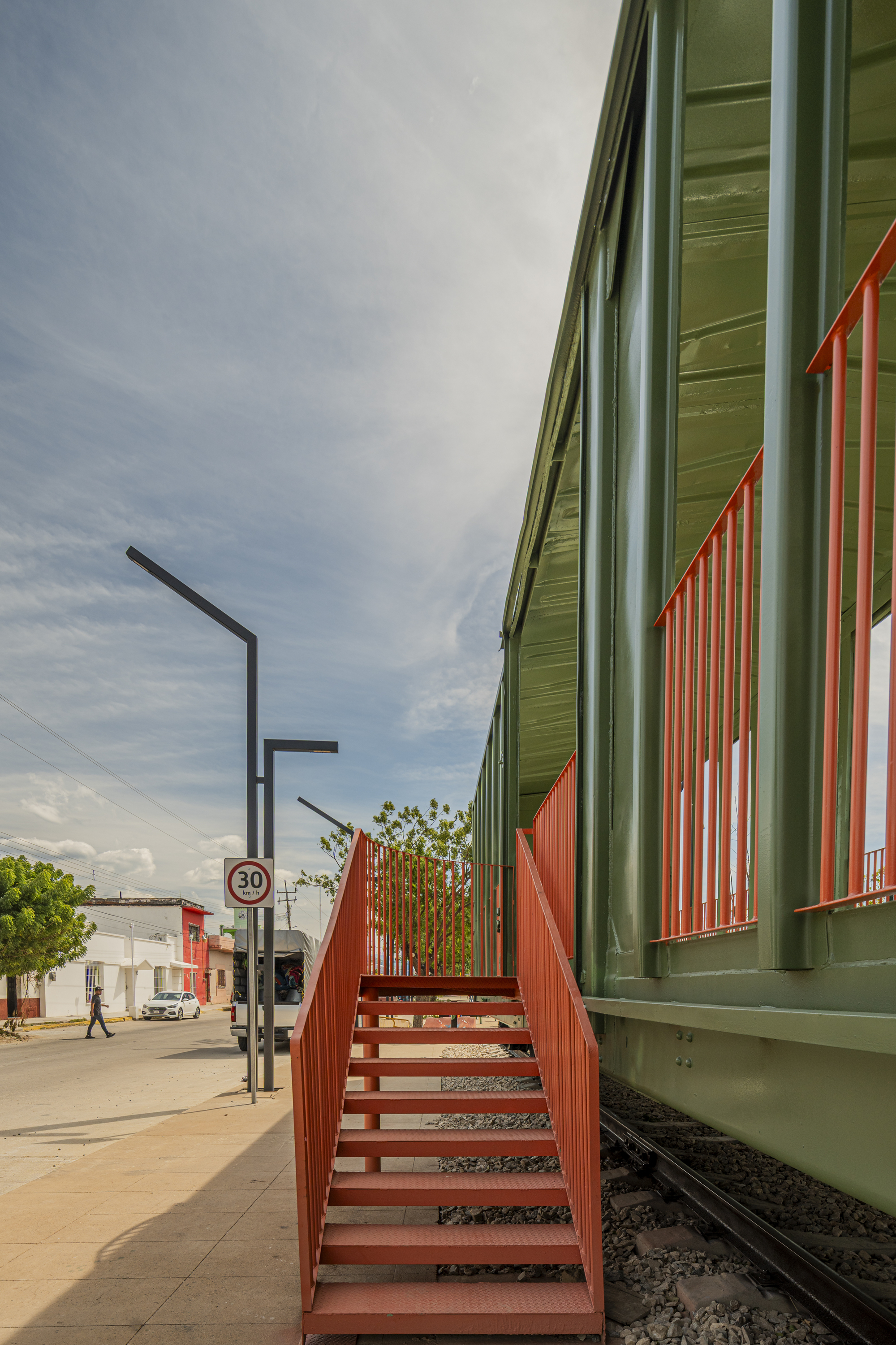 Orange steel stair rising to the green railcar, with a street, parked vehicle and lamp post beyond in afternoon light