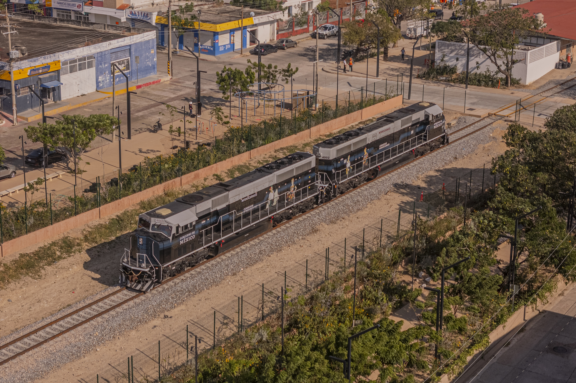 Aerial view of a blue and grey diesel locomotive pair moving along ballasted track beside a brick wall and street trees in afternoon sun