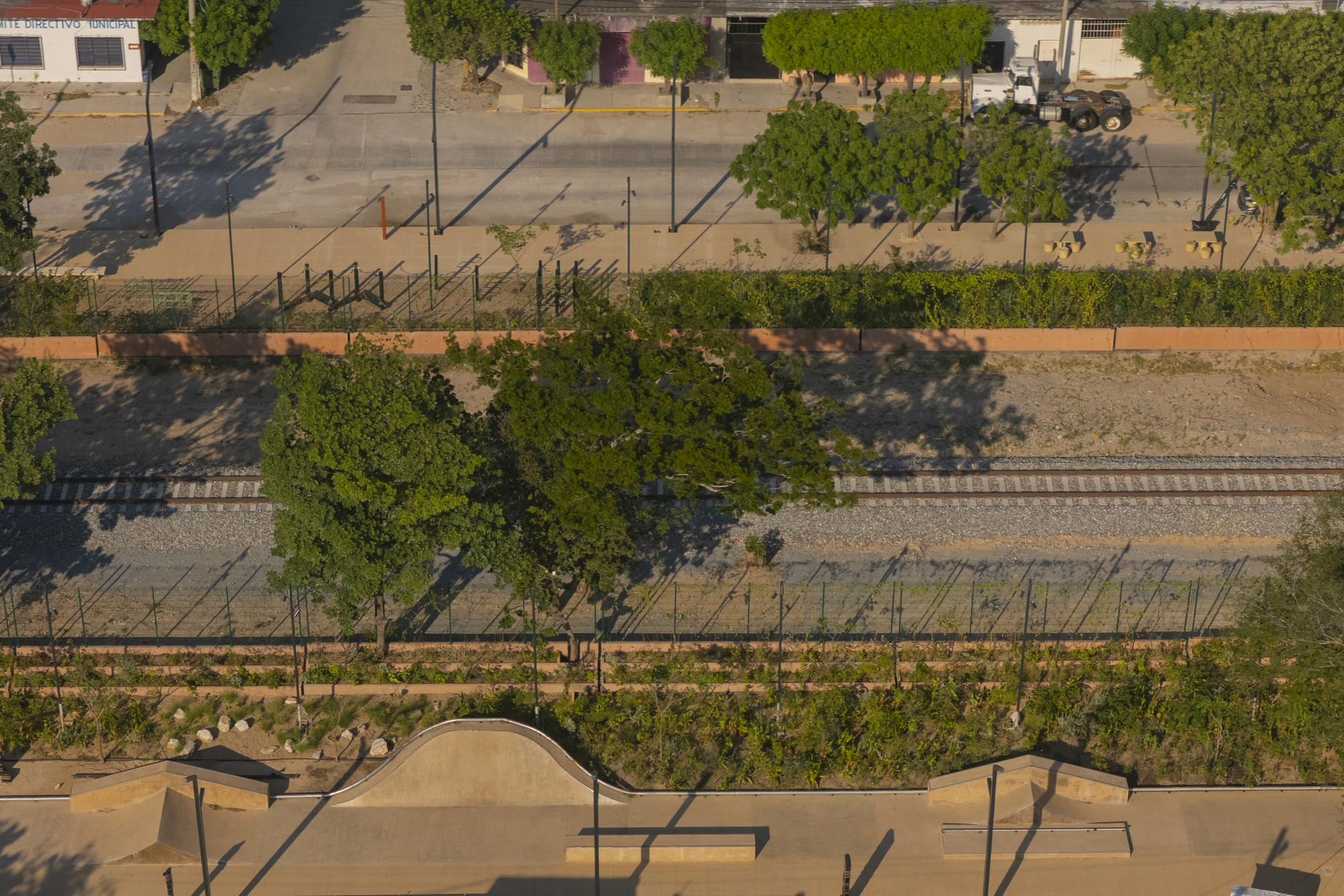 Top-down view of a planted strip, railway tracks and stone benches, with tree shadows falling across the gravel in low sun