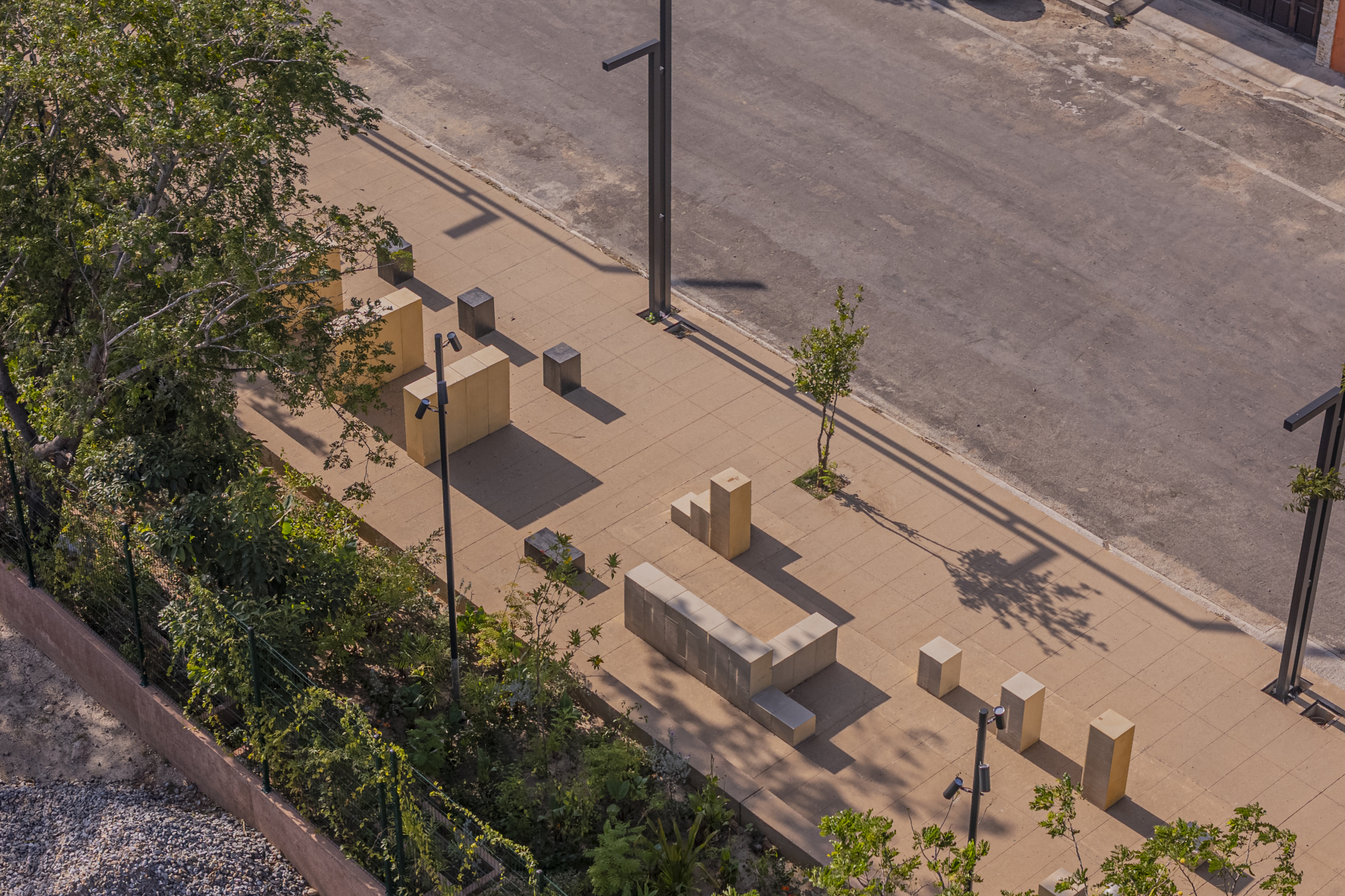 Aerial view of pale stone blocks and bollards scattered across a paved plaza, with long shadows from lamp posts and a young tree