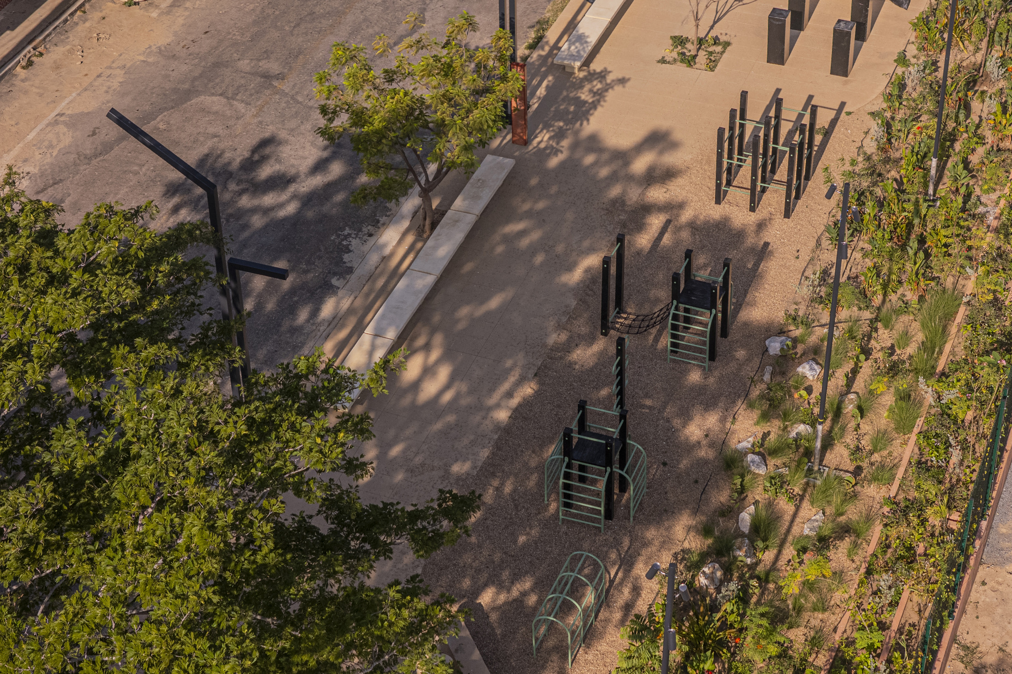 Overhead view of black and green exercise frames on a gravel court beside a planted slope, dappled by tree shadows