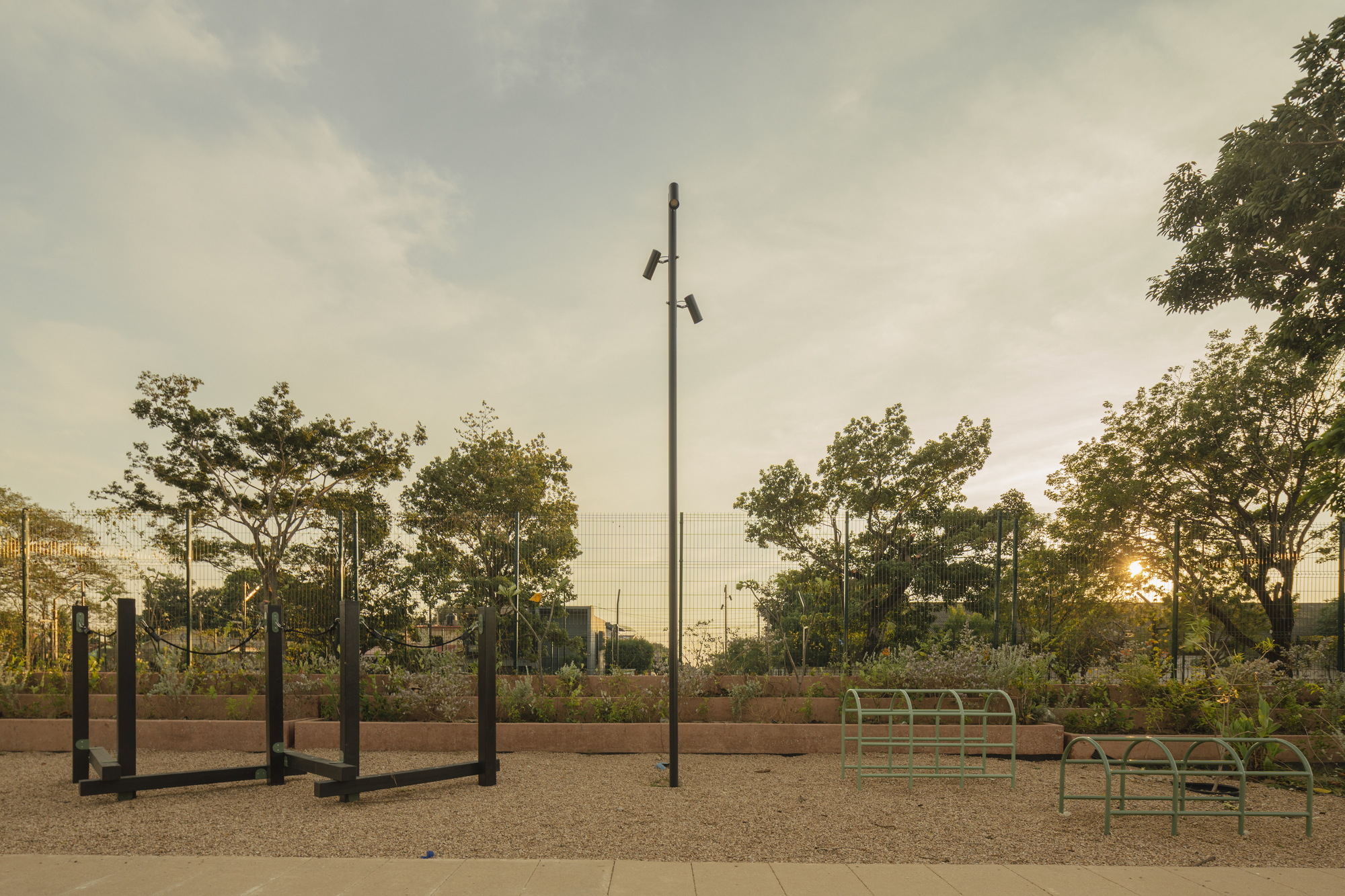 Black timber exercise bars and green metal racks on gravel before a planted terrace and mesh fence at sunset