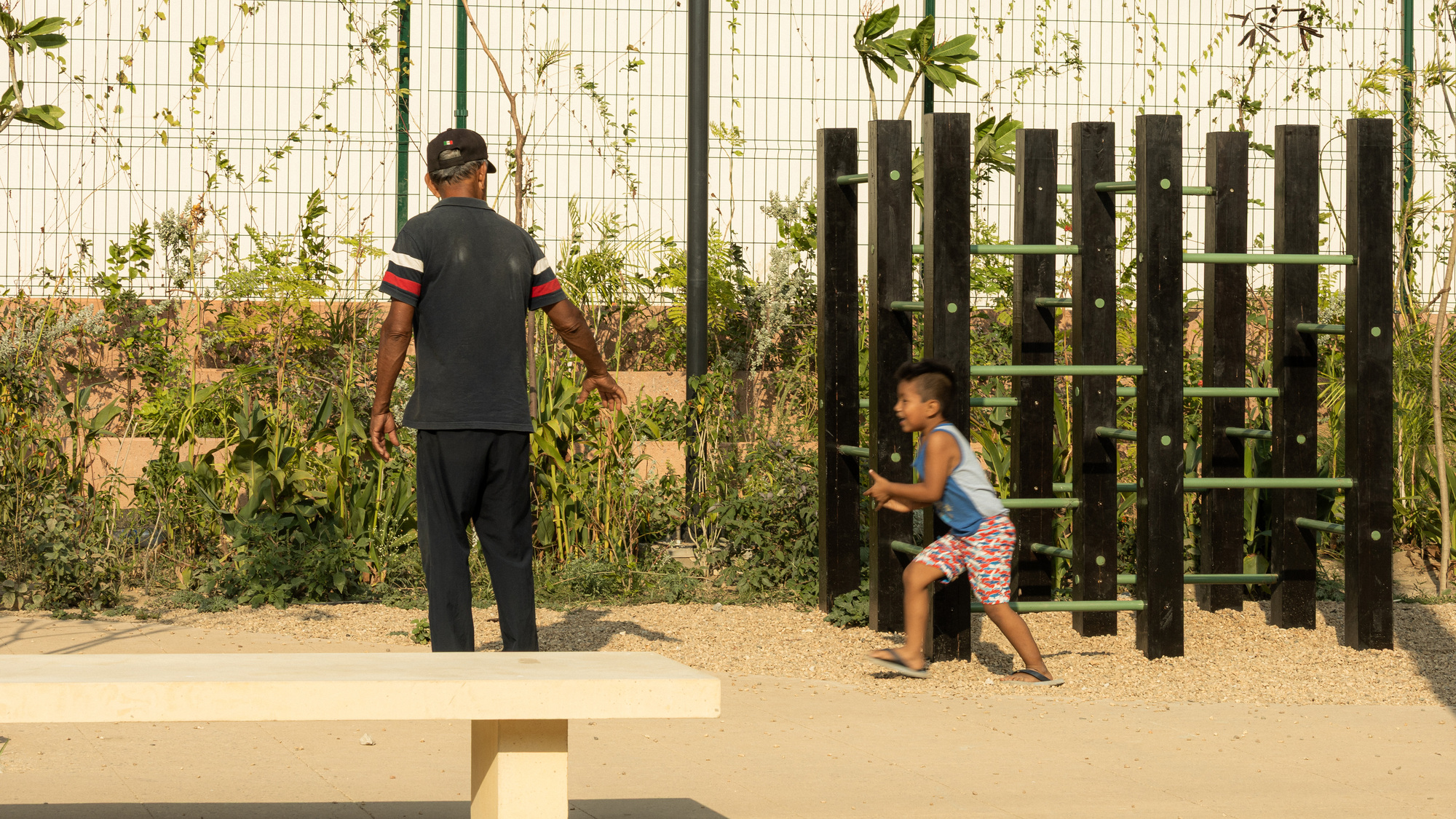 Boy running past a wall of black vertical posts with green rungs while a man stands near a concrete bench