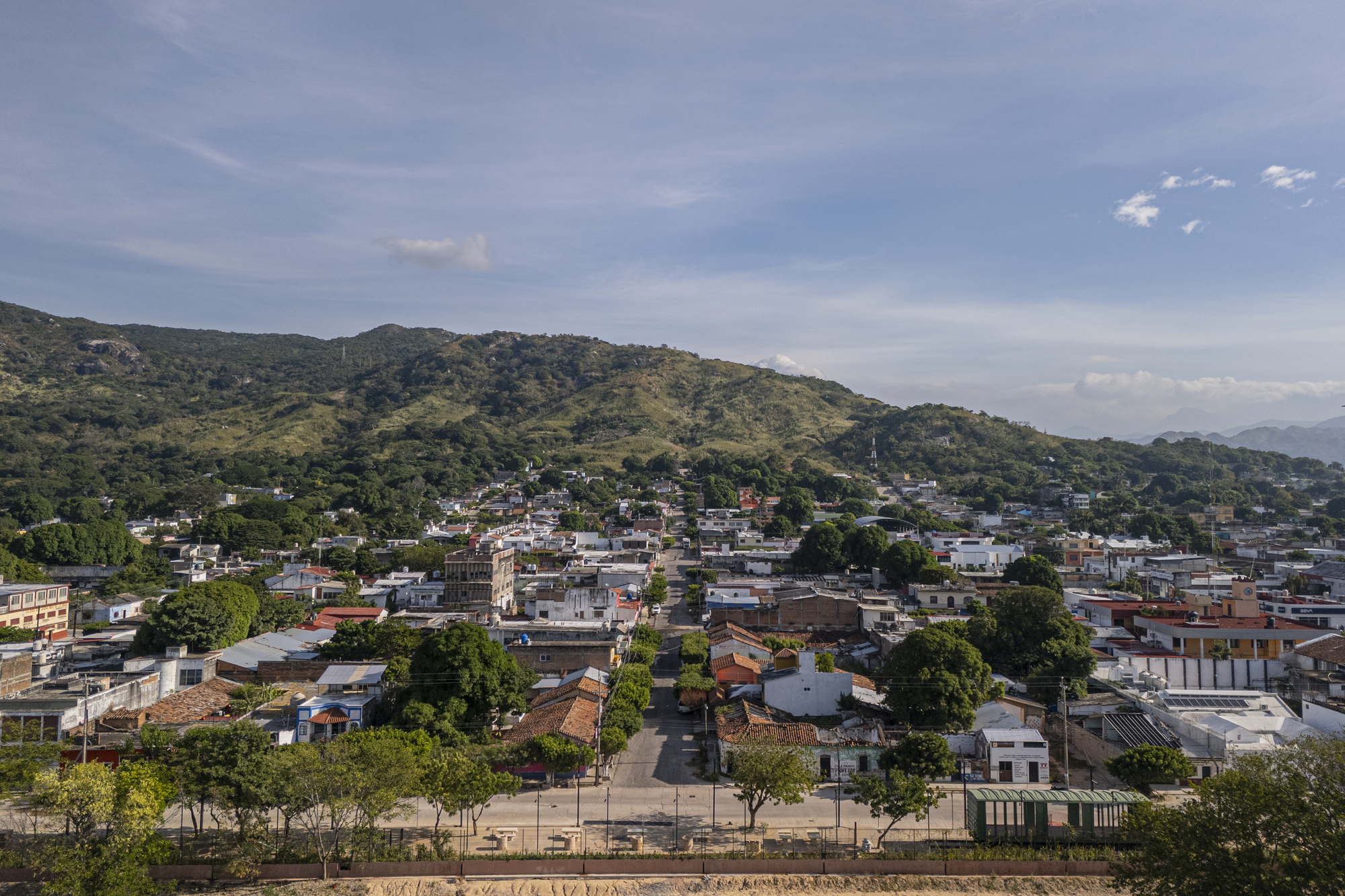 Aerial view of a low-rise town with tiled roofs and tree-lined streets below green hills under a cloud-streaked sky