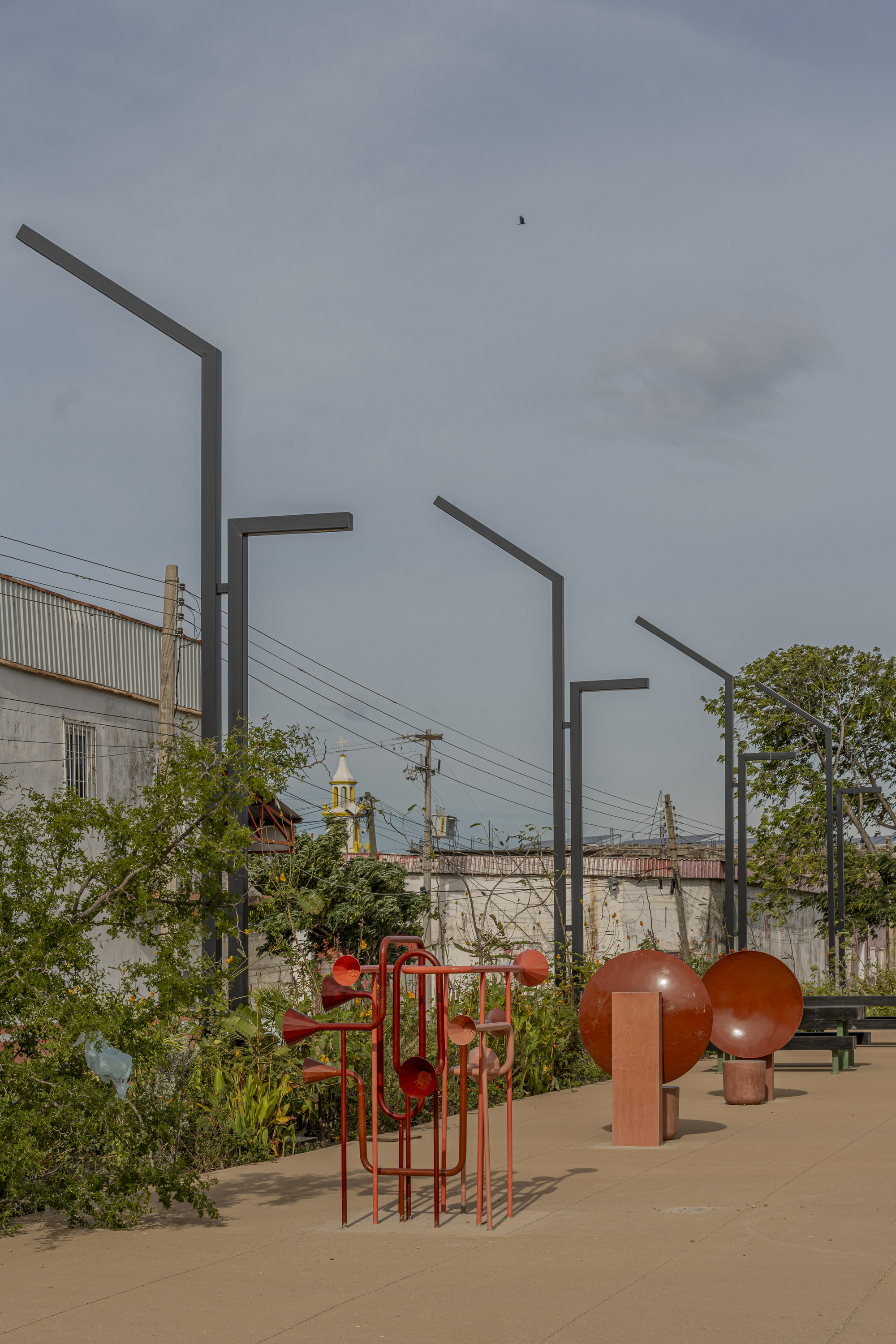 Red metal horn sculpture and round disc forms on a paved plaza beneath angled lamp posts and an overcast sky