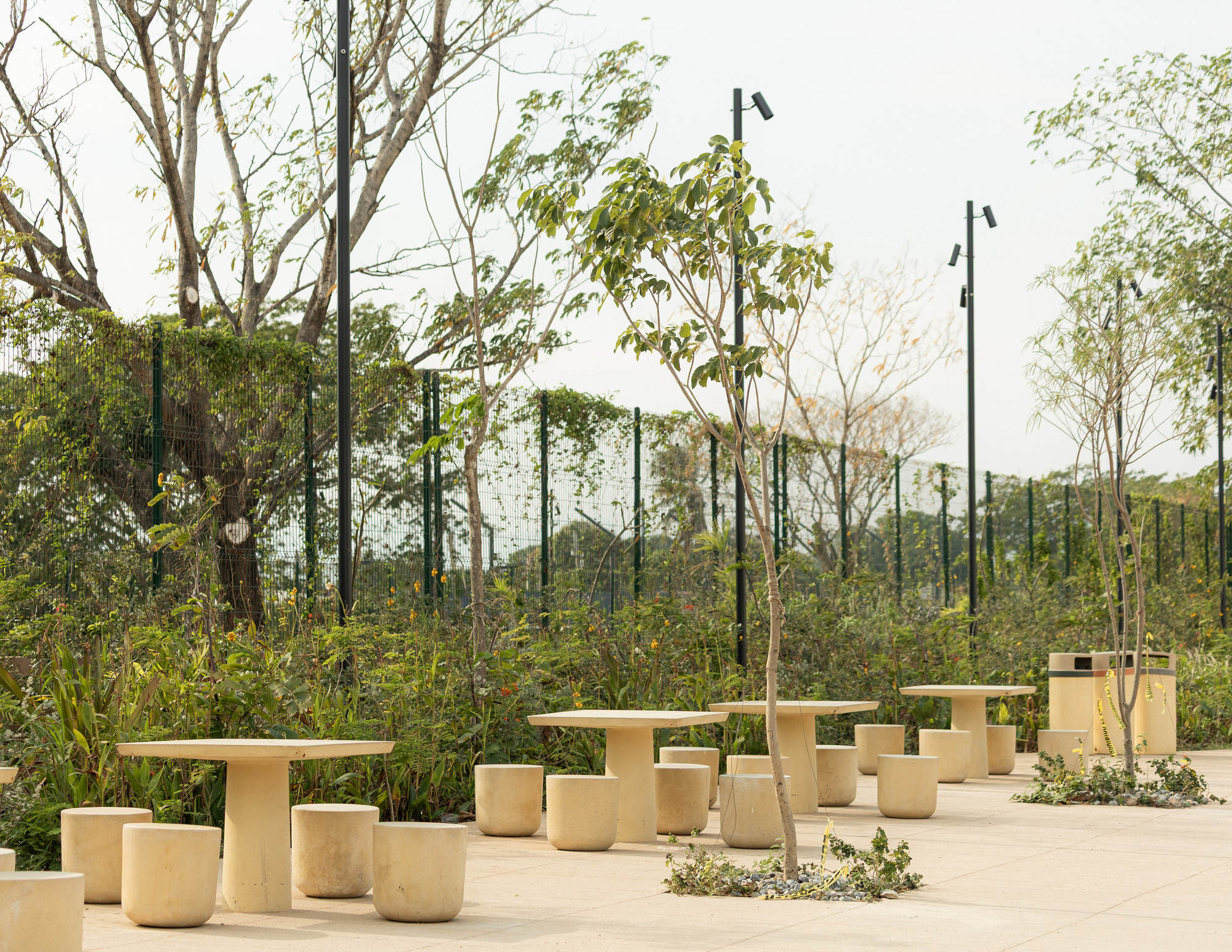 Round stone stools and pedestal tables on a pale paved square, with young trees, lamp posts and a green fence behind
