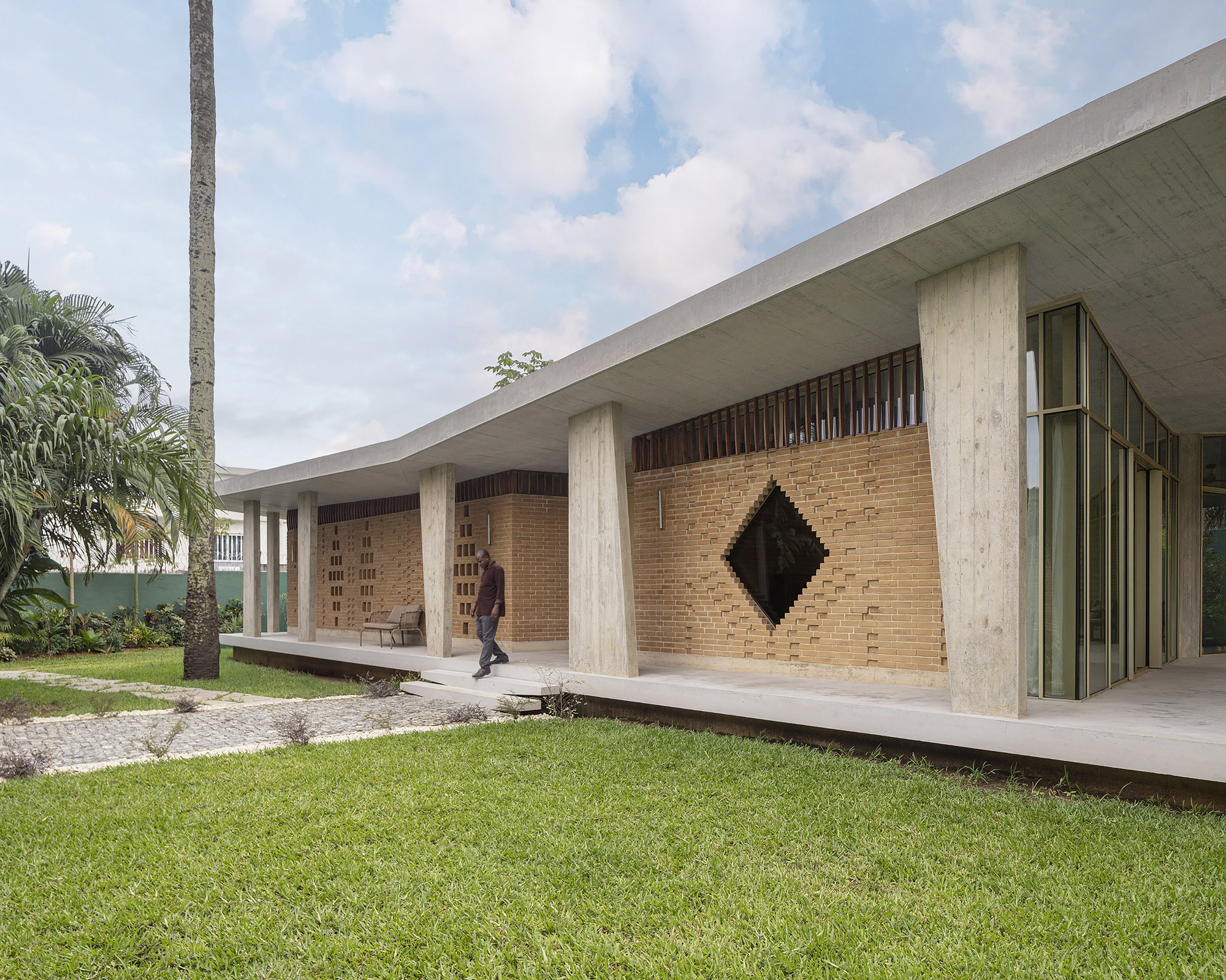 Brick wall with a diamond-shaped opening beneath a cantilevered concrete roof, a man walking along the veranda past a lawn and palm