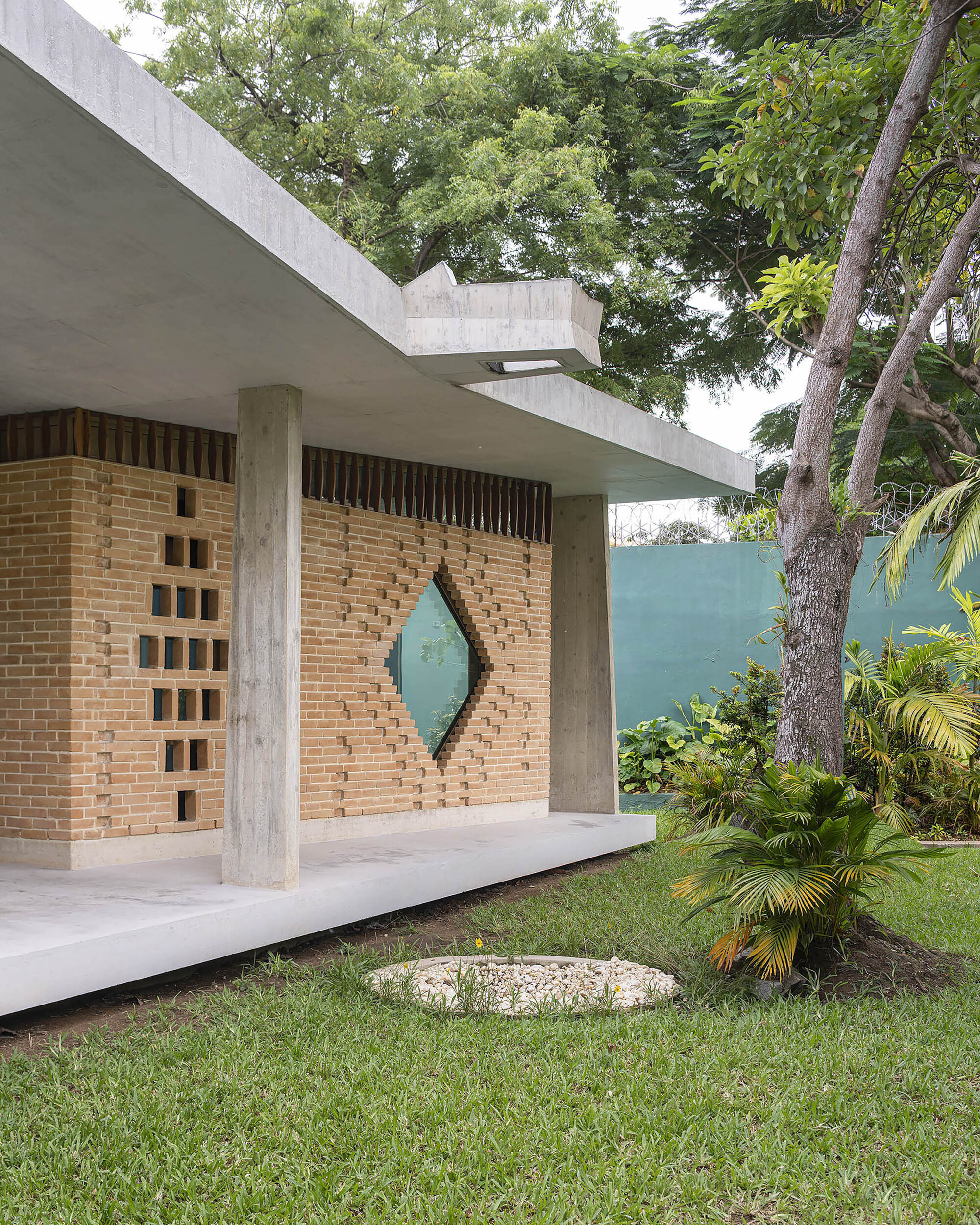 Corner of the brick wall with perforated openings and a diamond window, framed by concrete columns and a tree trunk