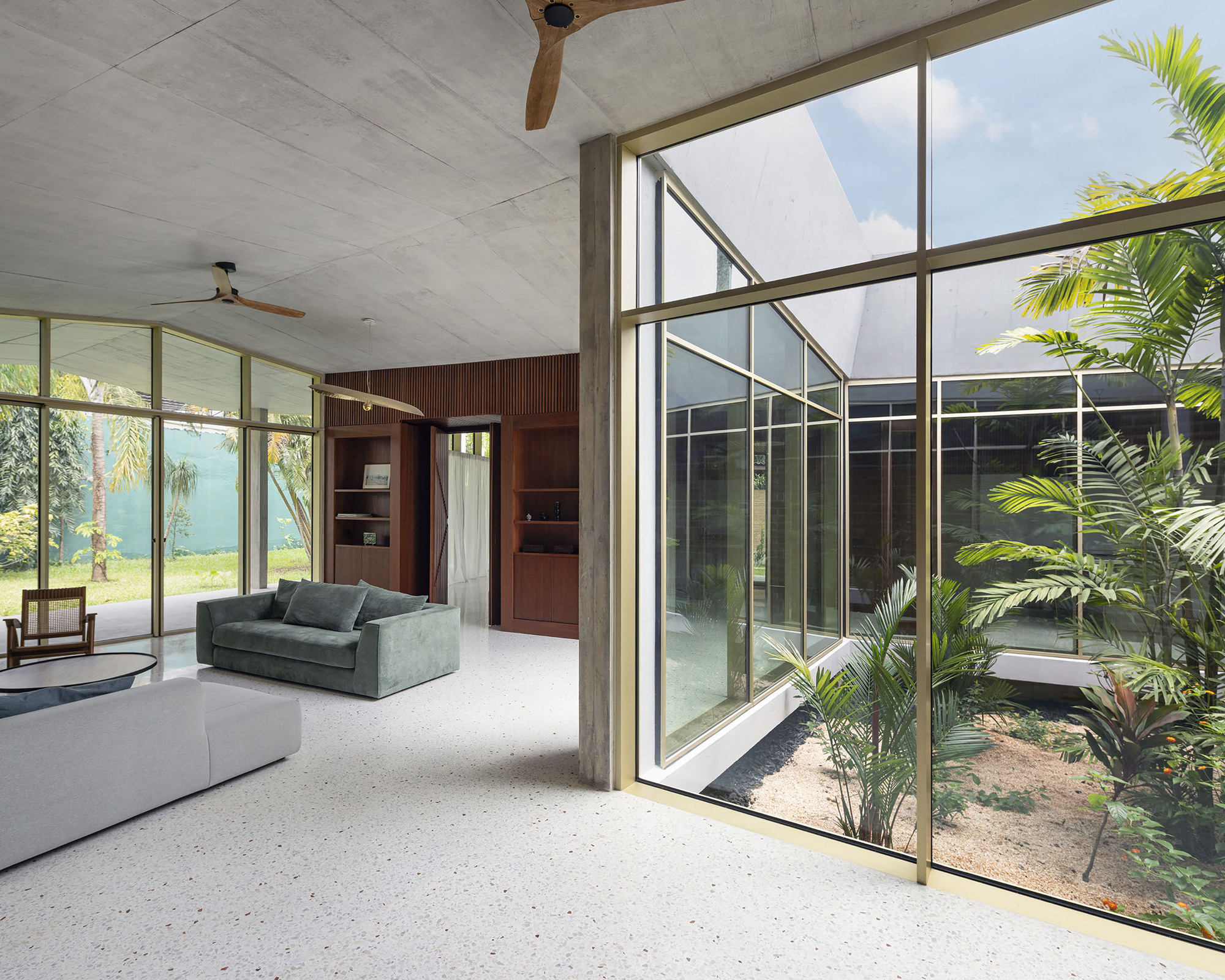 Living room with terrazzo floor and green sofa beside a glazed courtyard planted with palms, daylight through the glass