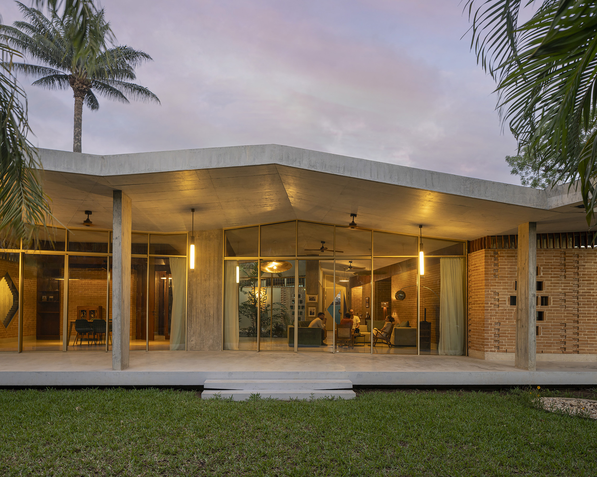 Front elevation at dusk with warm interior lights glowing behind full-height glazing under a folded concrete roof and palm trees