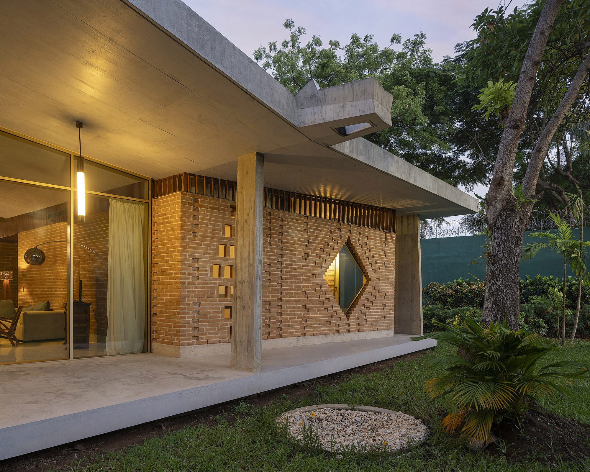Brick wall with diamond window and perforated openings at dusk, lit pendant lamp beside glazing and a tree on the lawn