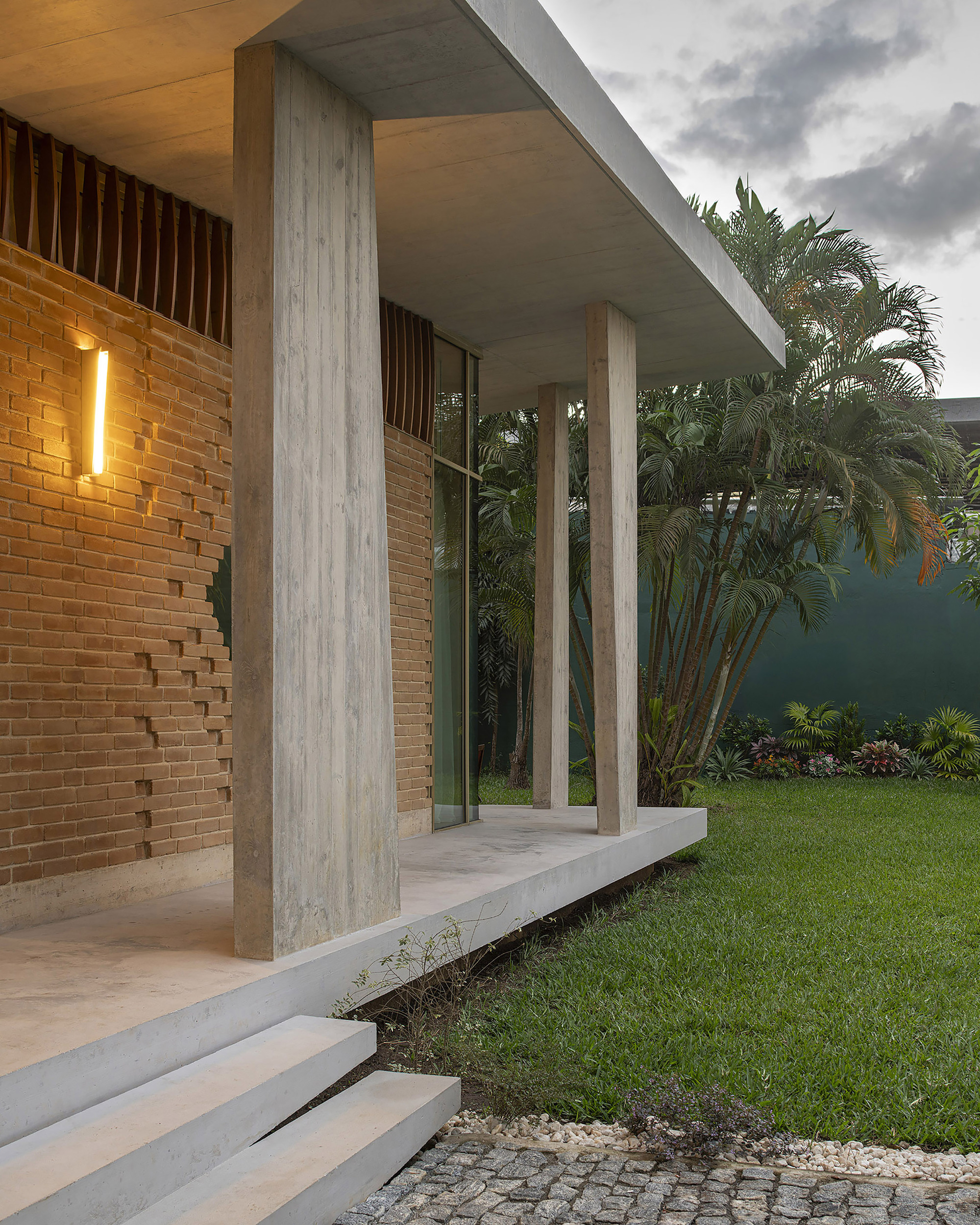 Board-formed concrete columns and flat roof slab over a brick wall with a wall lamp, beside a lawn at dusk