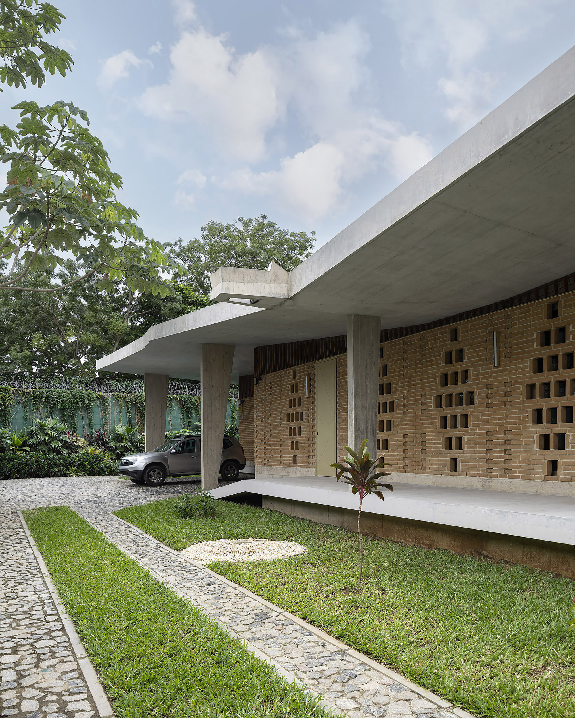 Cantilevered concrete roof over a perforated brick wall, with a parked car beyond a cobblestone path and lawn under cloudy sky