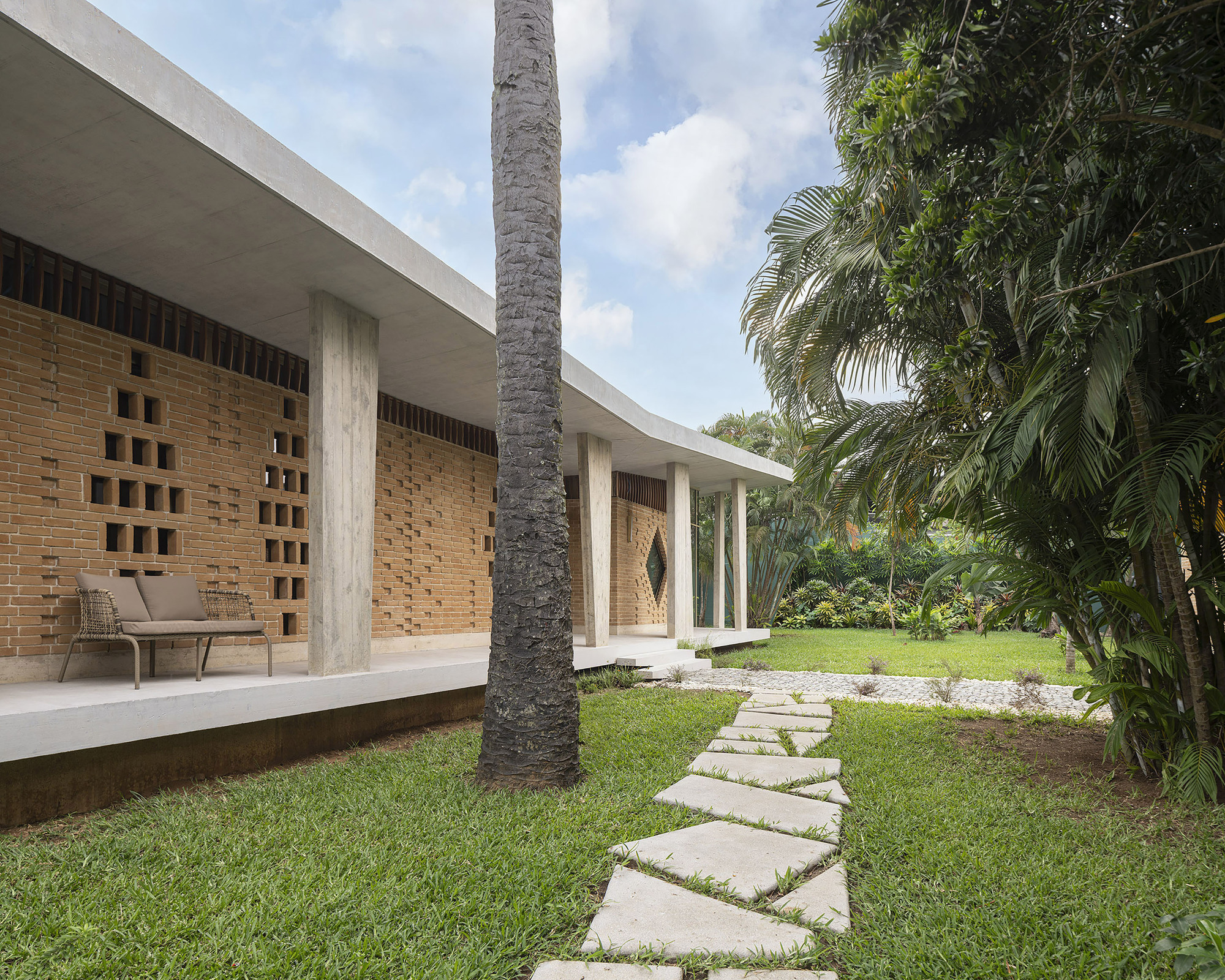 Covered porch with a wicker bench against a perforated brick wall, seen past a palm trunk and stepping stone path