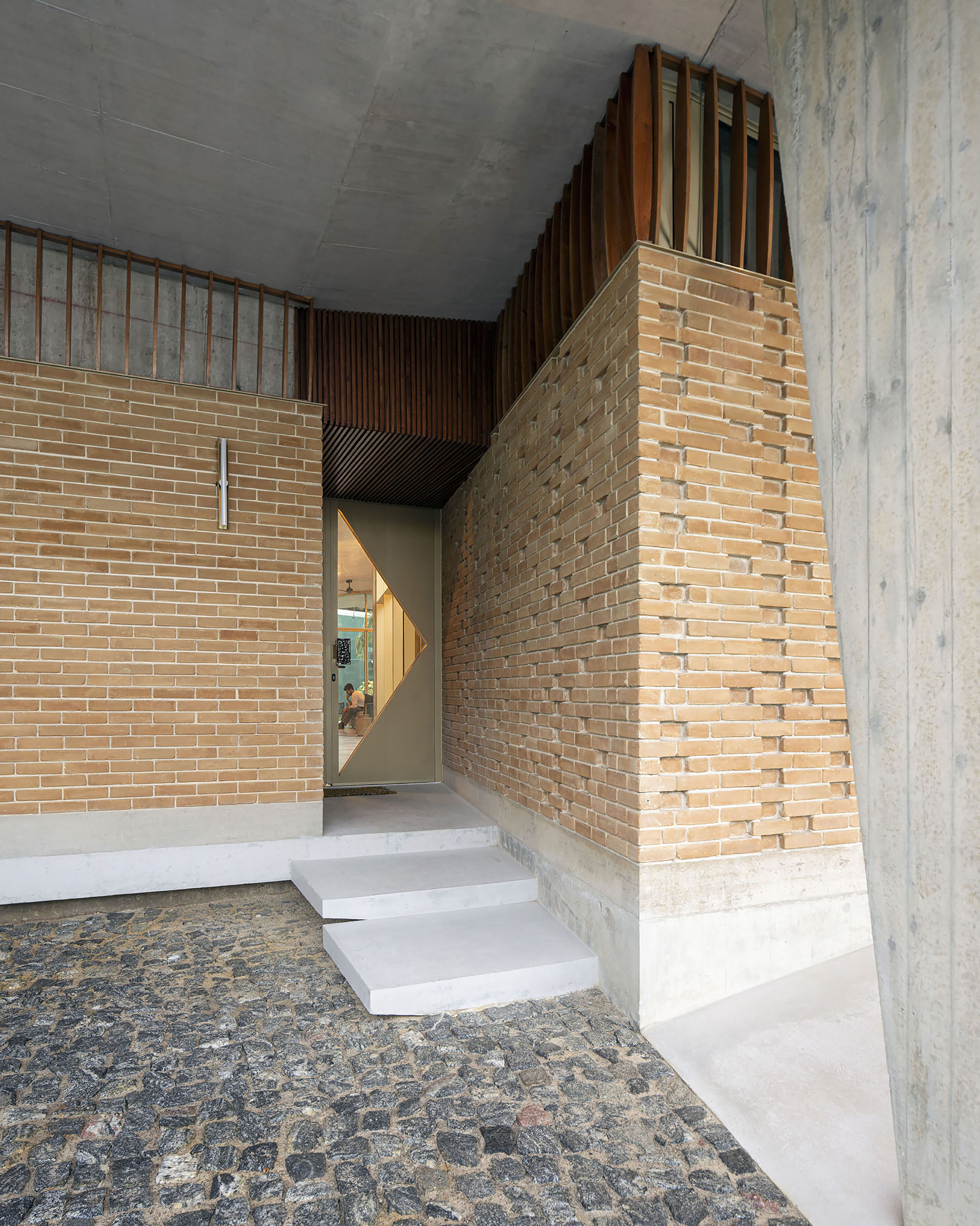 Entrance alcove with stepped concrete treads, a diamond-glazed door and brick walls topped with timber slats, over a cobblestone floor