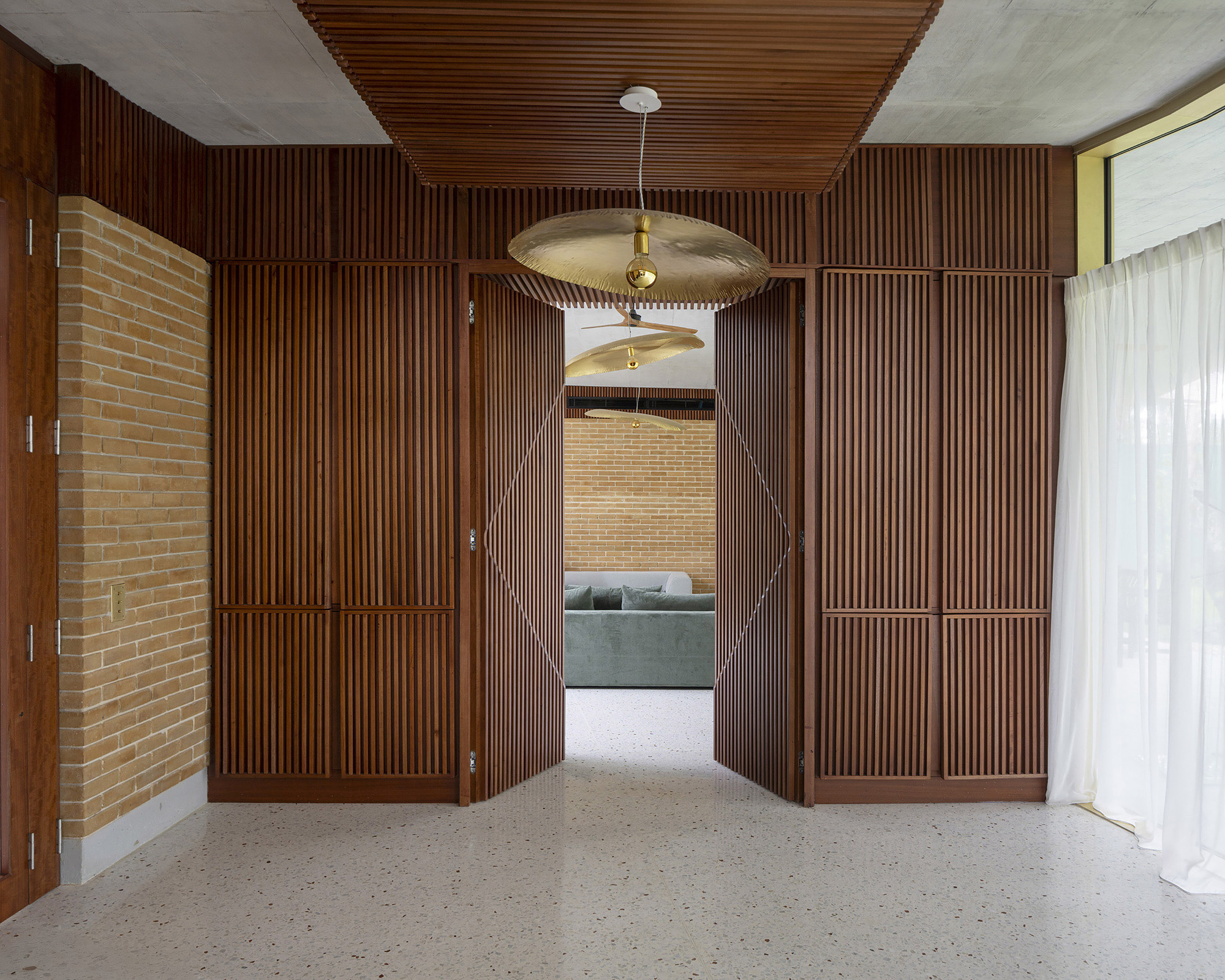 Timber slatted double doors opening from a terrazzo-floored room to a lounge, with a brass pendant lamp and sheer curtain