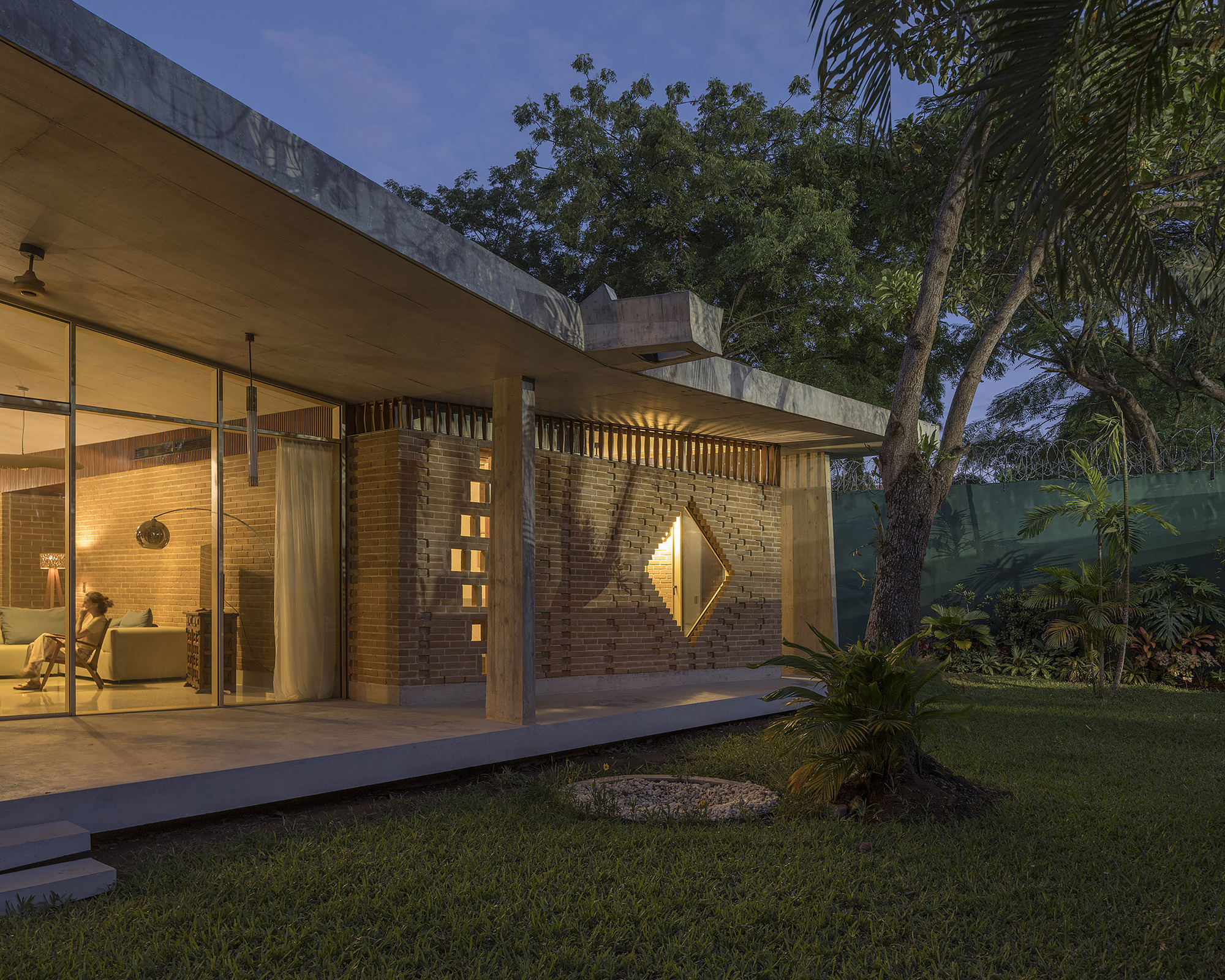 Dusk view of the glazed living room and diamond-windowed brick wall glowing under the concrete roof, with a seated person inside