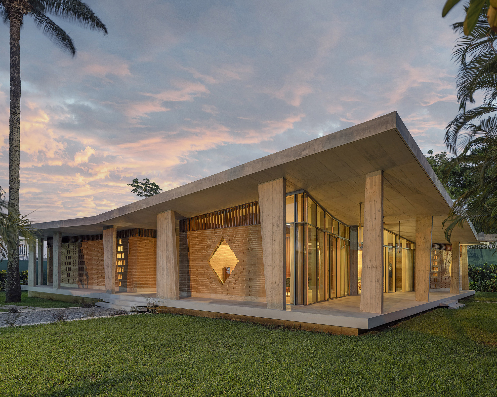 Corner view of the low brick pavilion with a diamond-shaped opening and tapered concrete columns under a pink dusk sky and palms