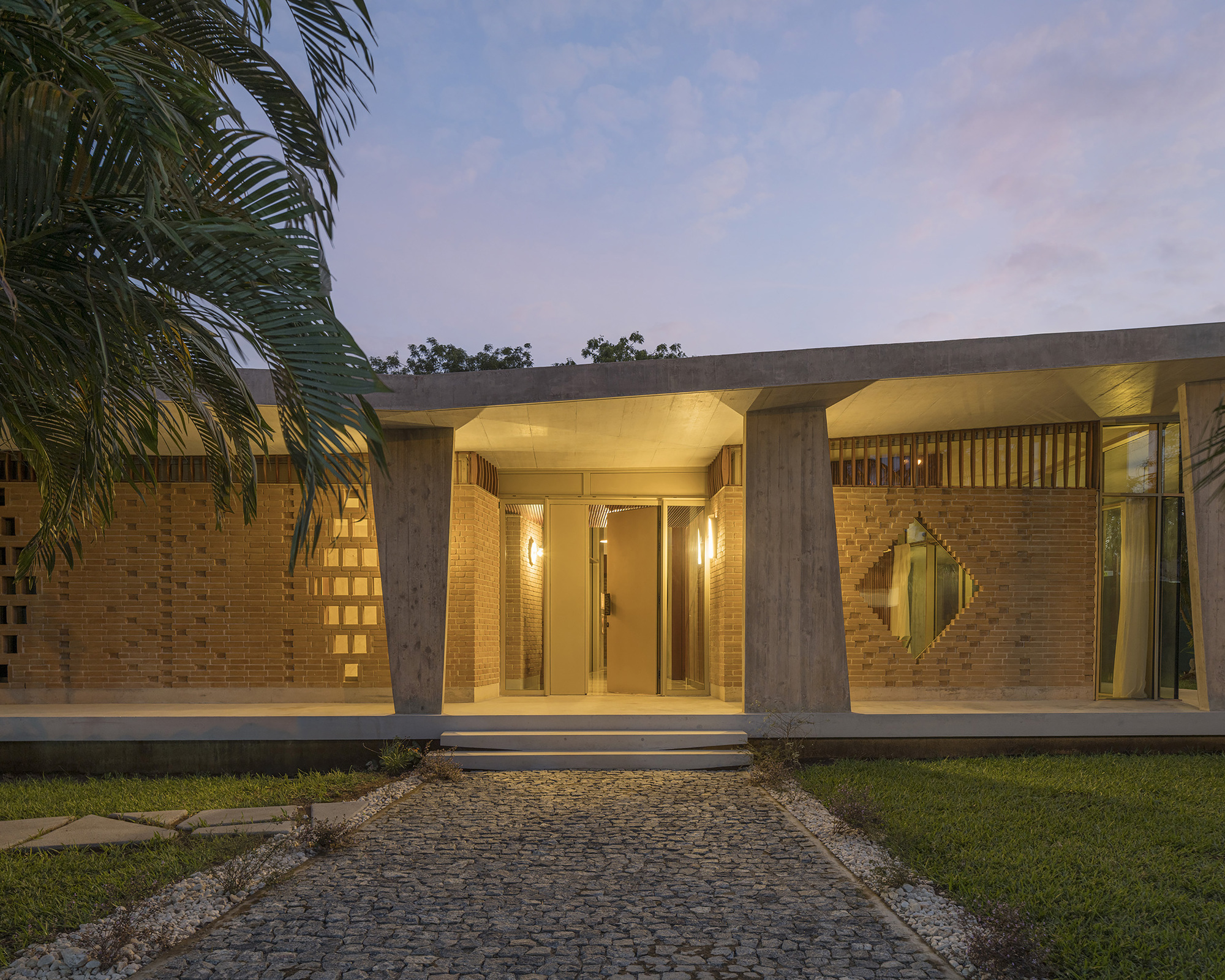 Entrance front with cobblestone path leading to a lit doorway between tapered concrete columns and brick walls at blue hour