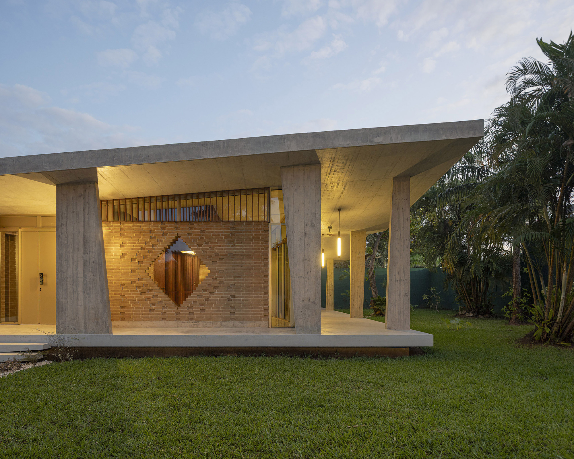 Brick wall with a diamond-shaped cutout beneath a thin concrete roof slab and slanted columns, lawn and palms at dusk