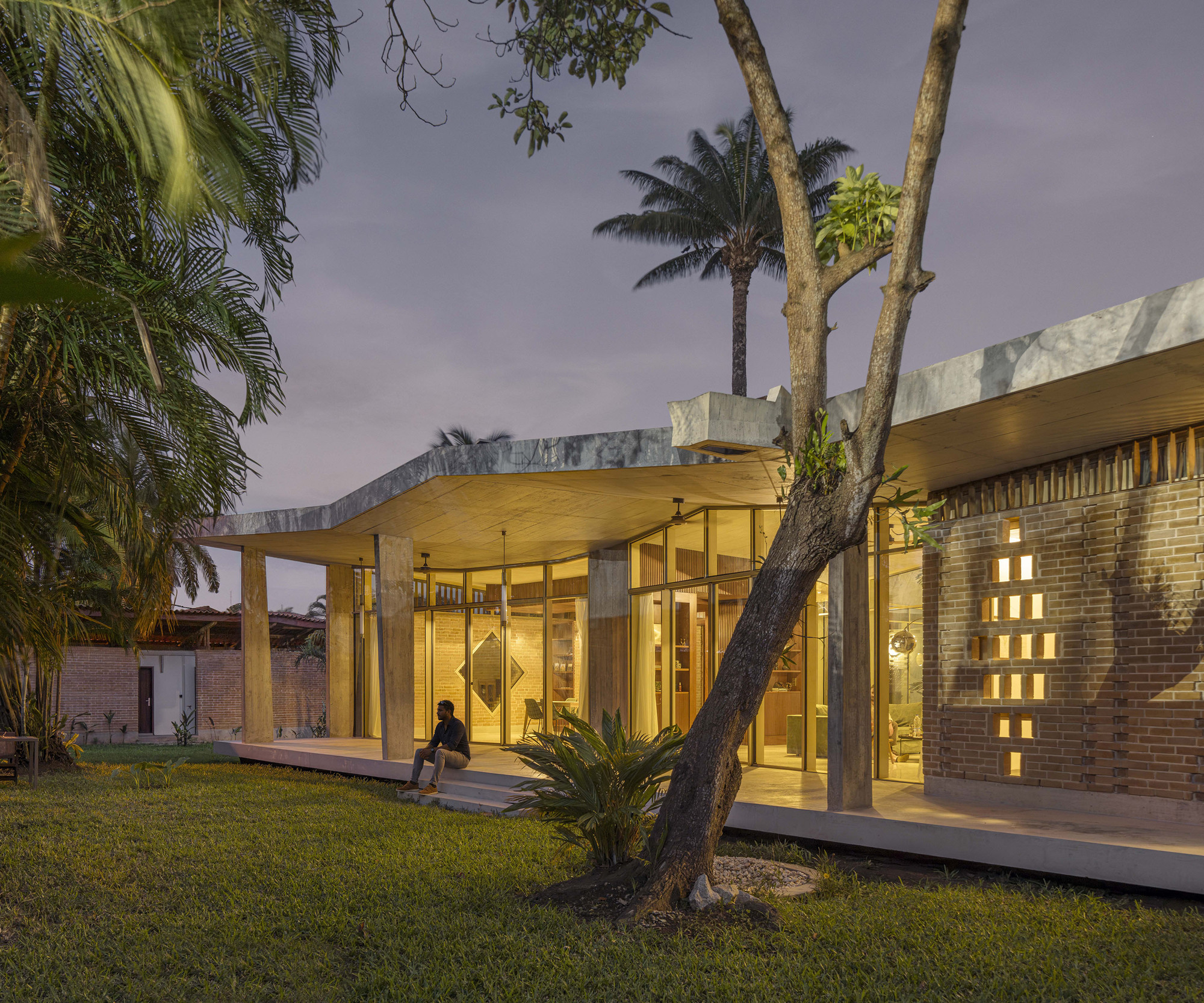 Veranda with glowing perforated brick screen and a seated person on the steps, a leaning tree trunk in the foreground at twilight